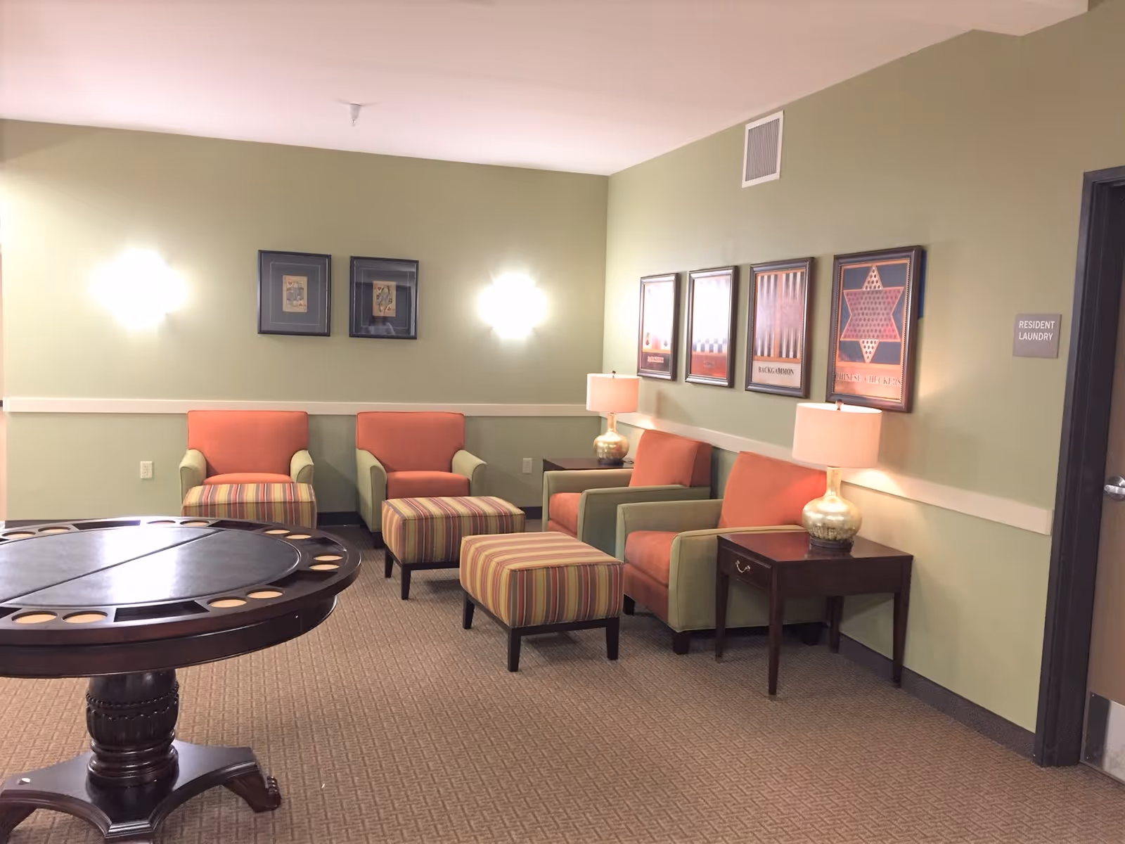 A cozy sitting area in a senior living facility with four armchairs upholstered in green and orange fabric, each paired with a striped ottoman. Two wooden side tables with lamps are placed between the chairs. The walls are painted light green and decorated with framed artwork. A round wooden game table is in the foreground. A door labeled 'Resident Laundry' is visible on the right side of the image.