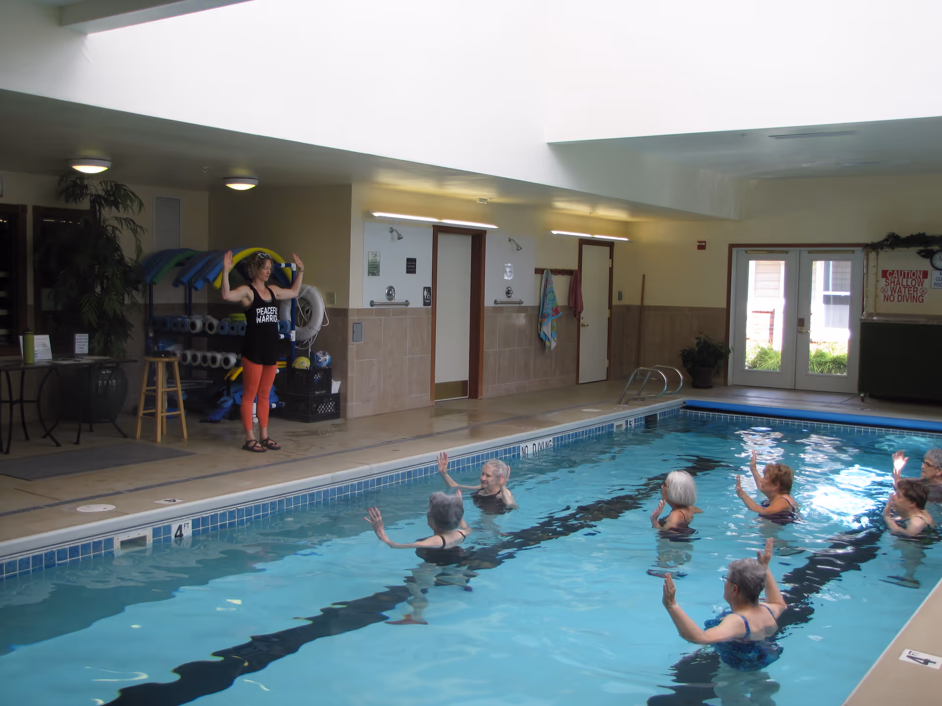 Indoor swimming pool with a group of elderly people participating in a water exercise class led by an instructor standing poolside. The instructor is wearing a black tank top and red leggings, demonstrating arm movements. The pool area has tiled walls, pool equipment, and double doors leading outside.