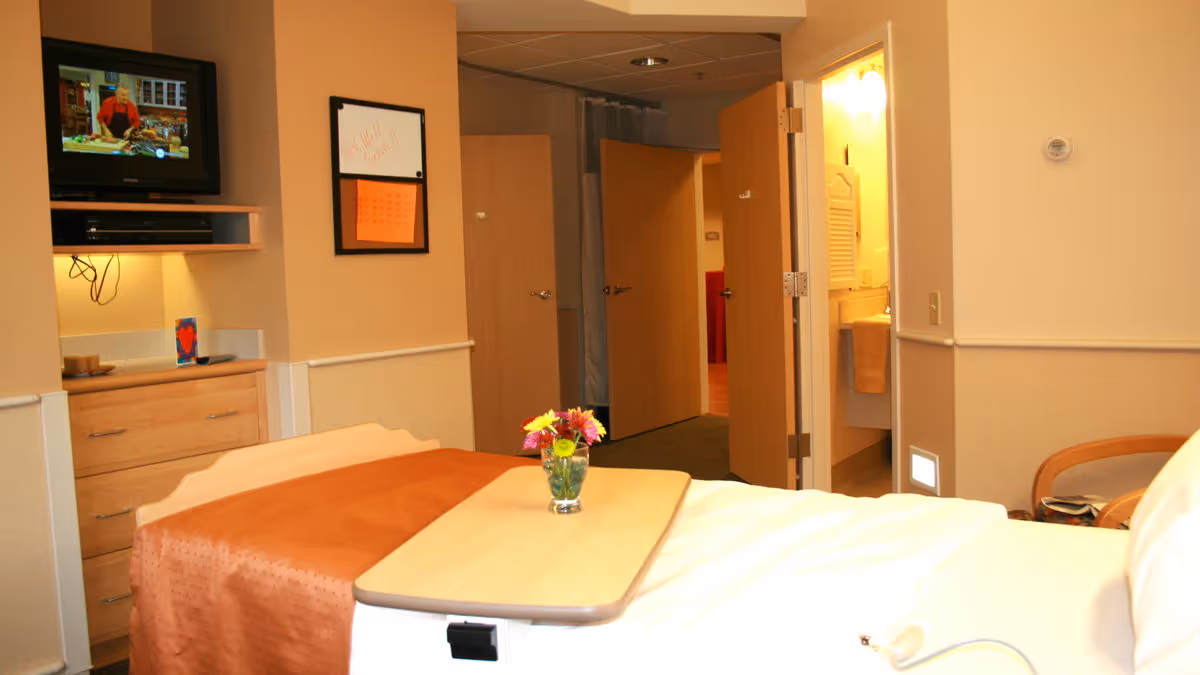 A senior living facility bedroom with a bed covered in white and orange bedding, a wooden overbed table with a small vase of colorful flowers, a wall-mounted TV showing a cooking show, a dresser, an open door leading to a bathroom with a sink and towel, and another open door leading to a hallway.