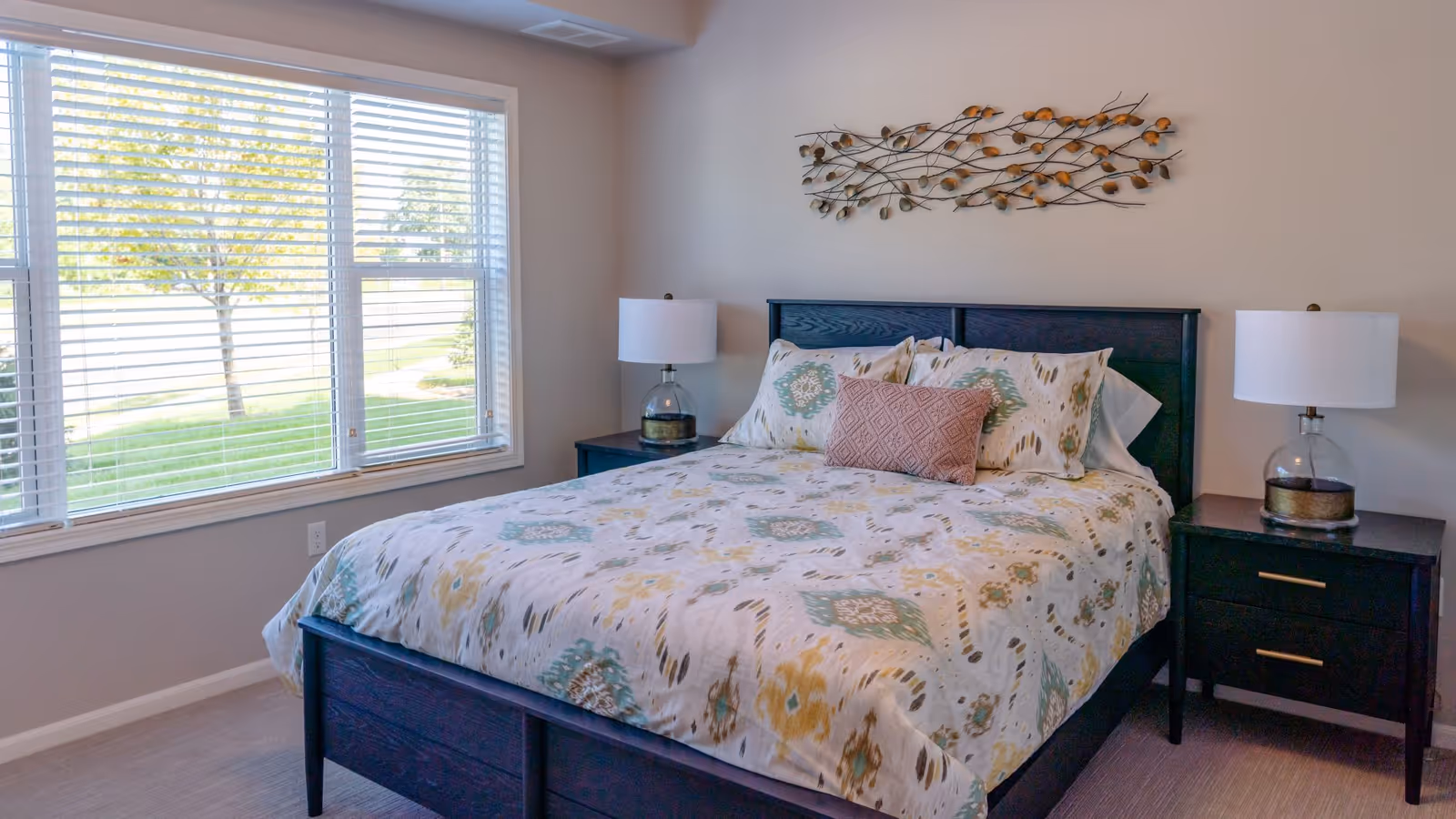 A bright bedroom with a large window showing green grass and trees outside. The room features a bed with a patterned comforter and pillows, flanked by two dark wooden nightstands each with a lamp. Above the bed is a decorative metal wall art piece with leaf designs.