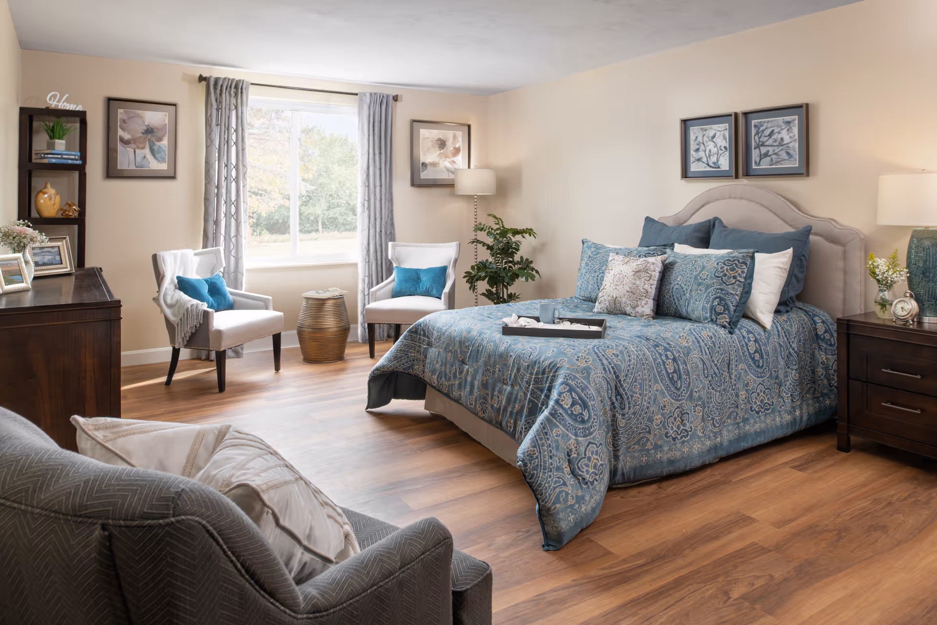 A well-lit bedroom with a large bed covered in a blue patterned comforter and multiple pillows. There are two white armchairs with blue cushions near a window with gray curtains. The room has wooden flooring, a dark wooden dresser, a small round side table, a potted plant, and framed artwork on the walls. A cozy armchair with pillows is in the foreground.