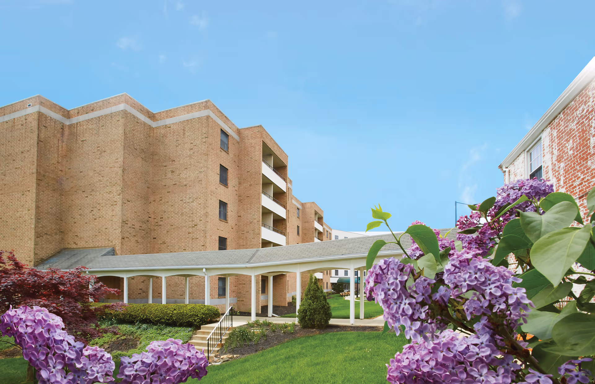 Brick senior living building with a covered walkway and purple flowers in the foreground under a blue sky.