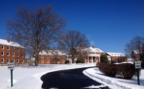 Snow-covered driveway leading to a large brick building with white columns and multiple windows under a clear blue sky. Leafless trees and trimmed bushes line the driveway, with 'EXIT ONLY' signs on both sides of the road.