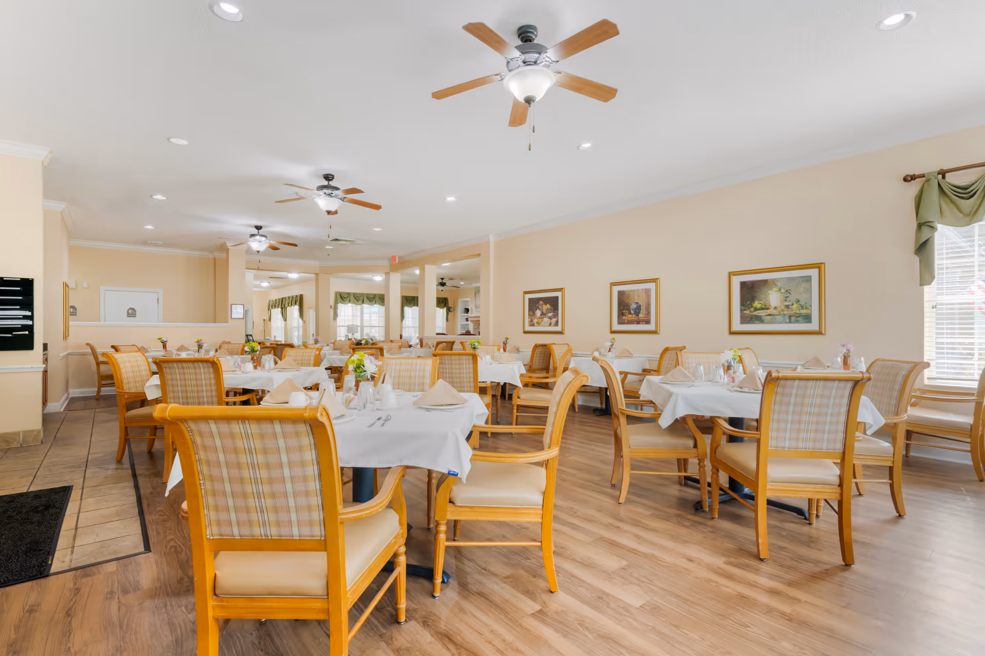 Bright dining room with multiple round tables, white tablecloths, and plaid-backed wooden chairs.