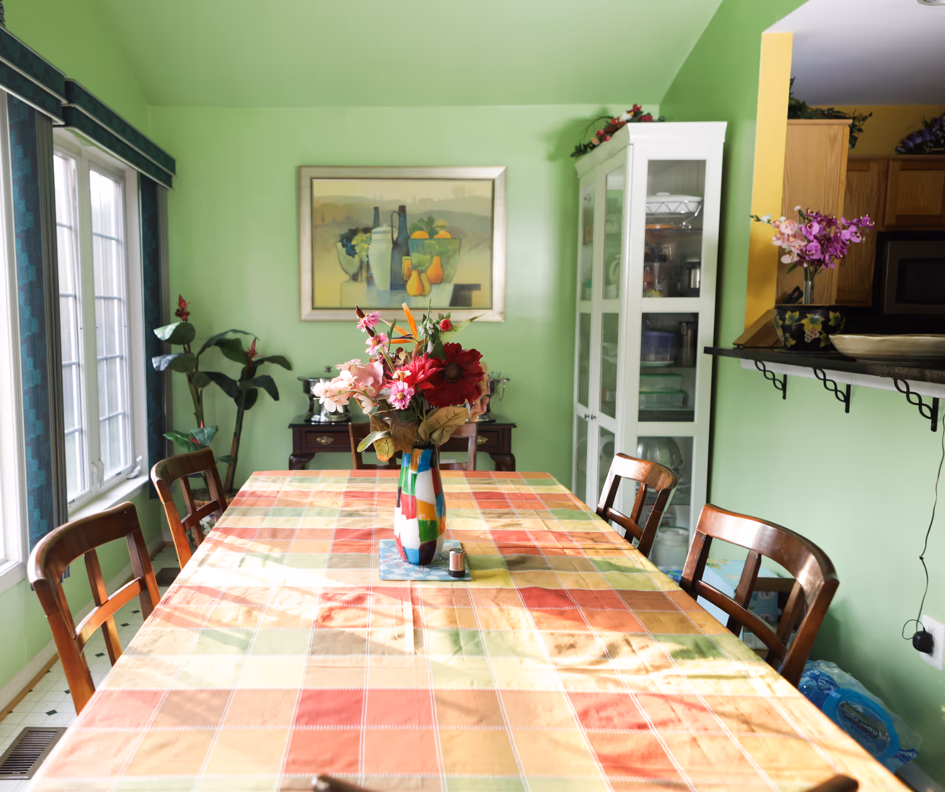 A dining room with a rectangular table covered with a colorful checkered tablecloth. There is a vase with a bouquet of flowers in the center of the table. Four wooden chairs surround the table. The walls are painted light green, and there is a large window with blue curtains on the left. A white cabinet with glass doors and a framed painting of fruit are visible on the far wall. To the right, there is a partial view of a kitchen area with wooden cabinets and a counter with a flower vase.