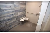 Accessible shower area with a fold-down white bench attached to the wall, a handheld showerhead, and a horizontal grab bar. The shower walls have a wood-like textured panel on one side and a plain light-colored wall on the other. The floor is a dark, textured surface designed to prevent slipping.
