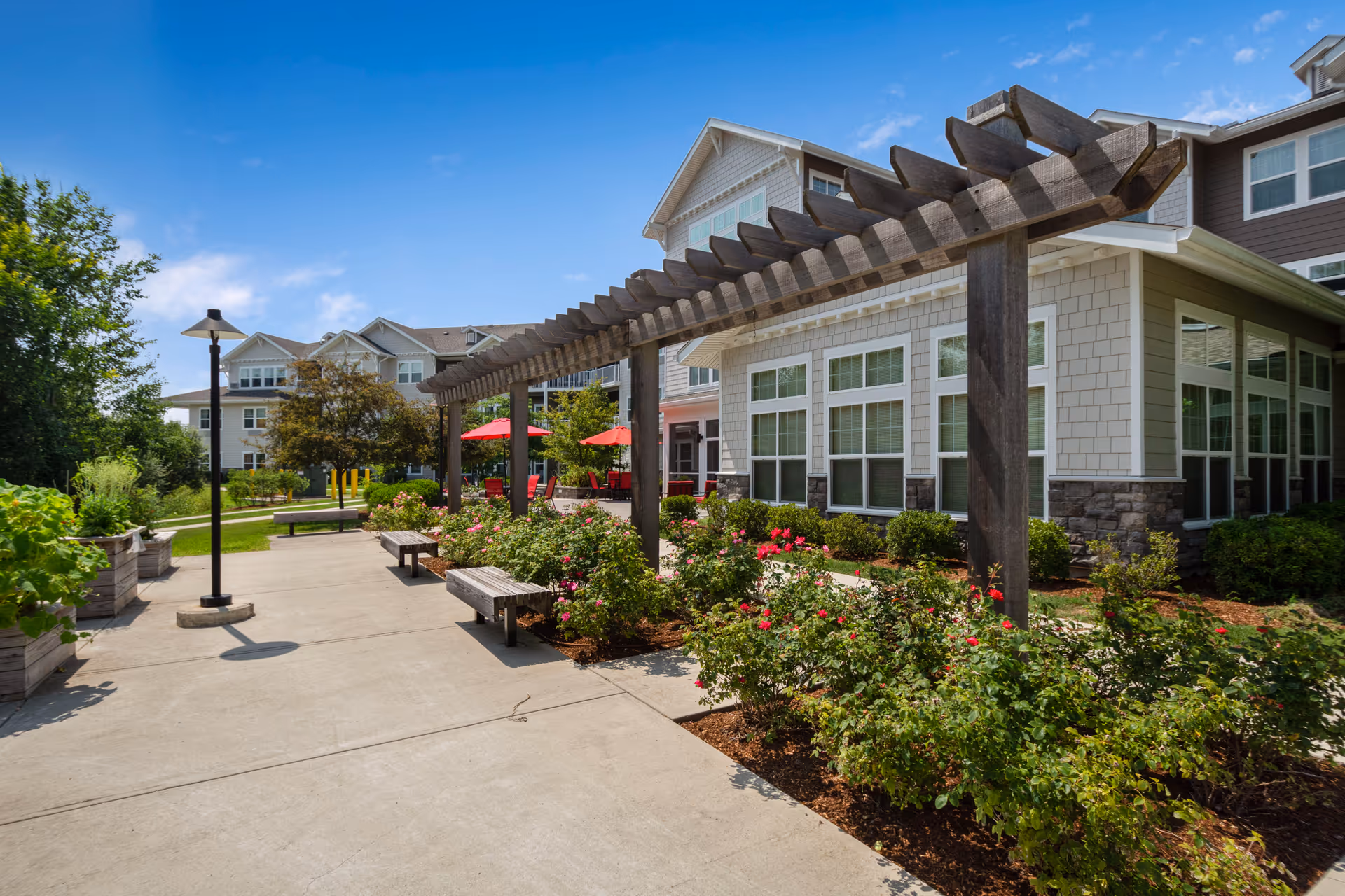 Outdoor courtyard with a wooden pergola, benches, flowerbeds and a multi-story retirement building in the background.
