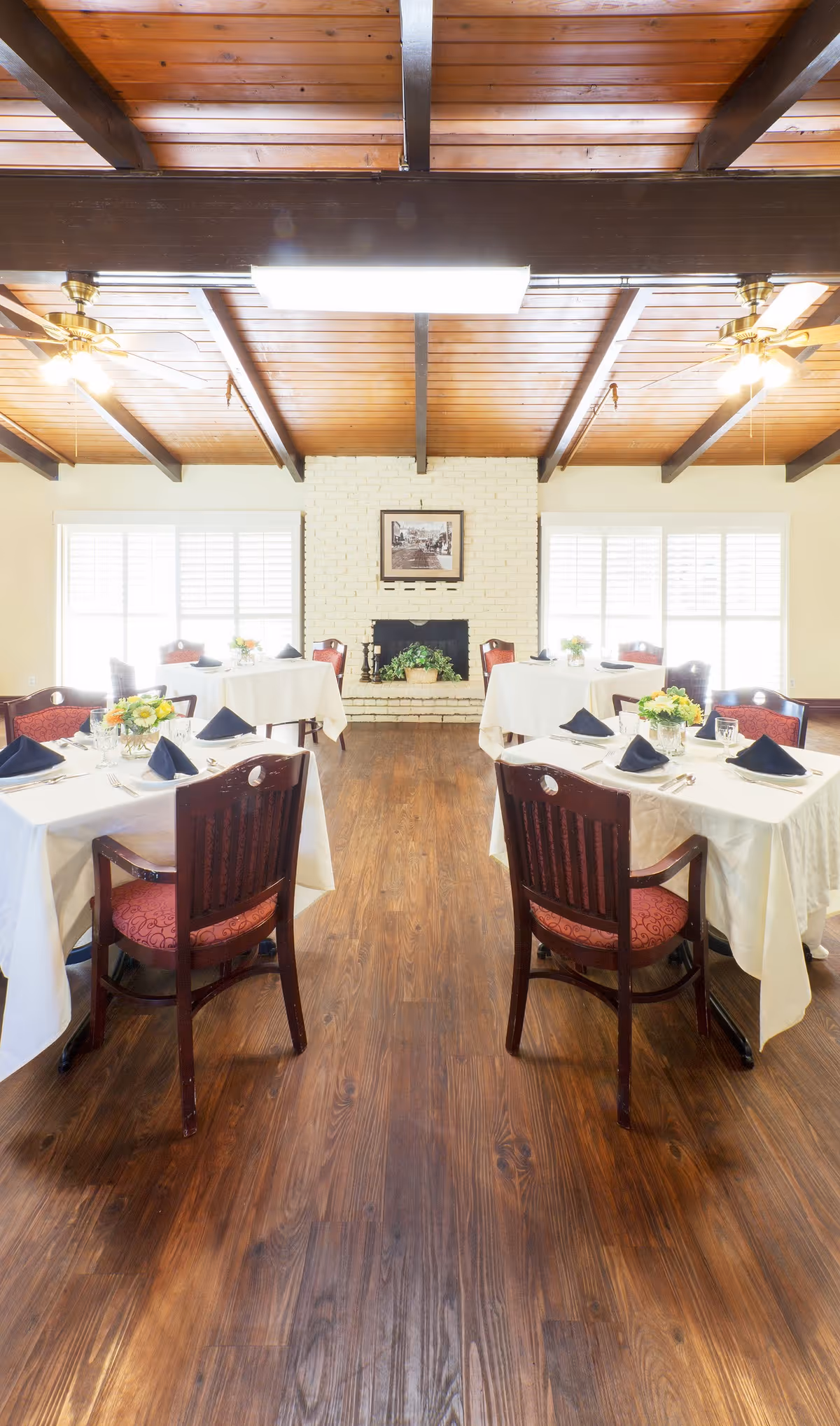 A bright dining room with wooden floors and a wooden beam ceiling. There are four tables covered with white tablecloths, each set with plates, glasses, silverware, and navy blue napkins. Each table has a small floral centerpiece. The room features a white brick fireplace with a framed picture above it and large windows with white shutters on either side.