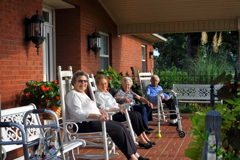 Four elderly women sitting on rocking chairs on a covered porch of a red brick building, with plants and greenery around them. Two walkers are visible nearby.
