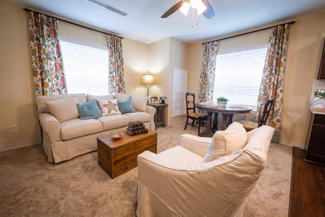 Cozy living room with two cream sofas, a wooden coffee table, and a small round dining table by windows with patterned curtains.