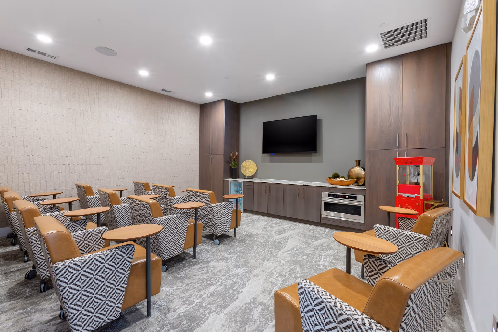 Small screening room with rows of patterned armchairs with tray tables facing a wall-mounted TV and built-in cabinetry, with a popcorn machine at the side.