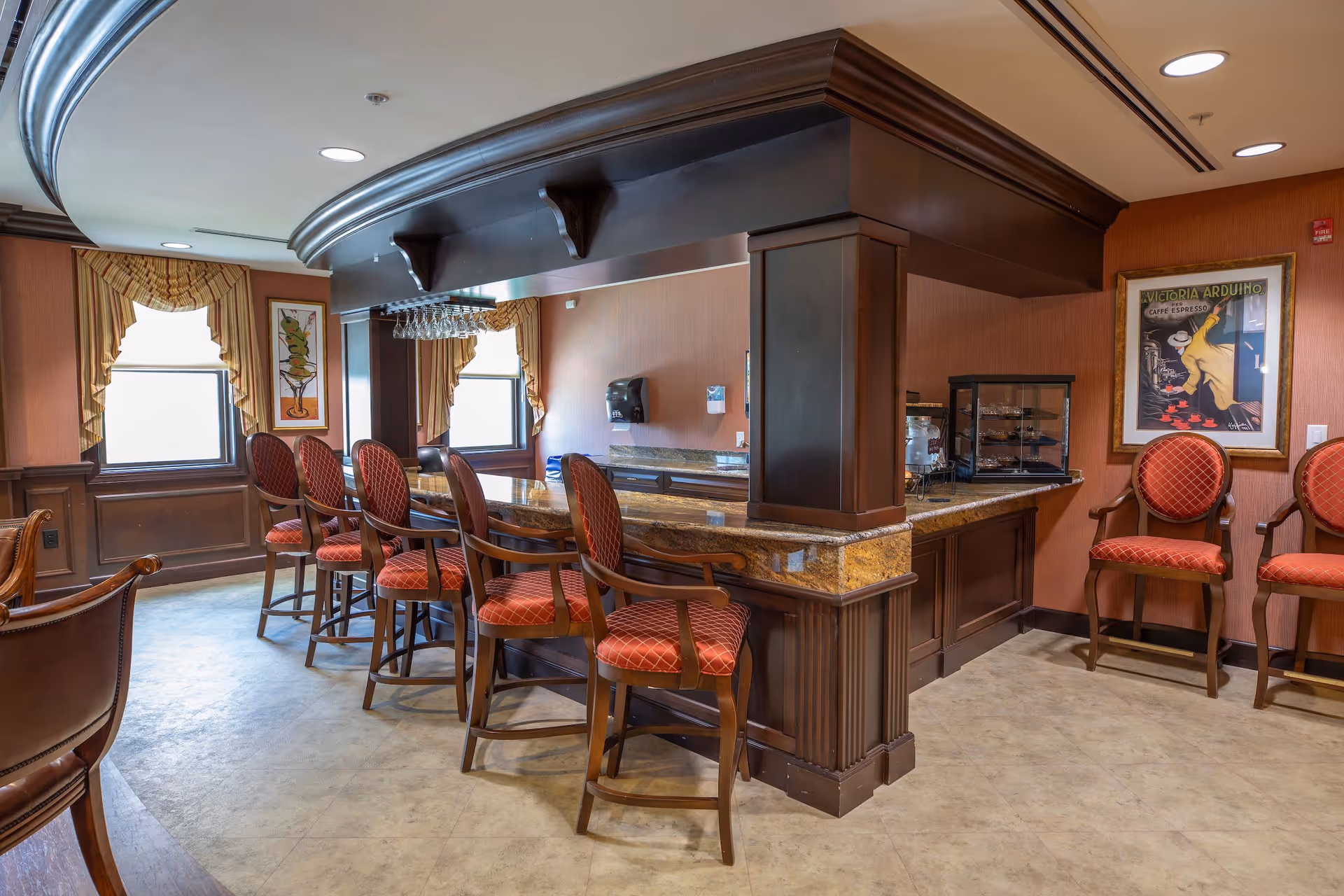 Interior lounge with a wood and marble bar, high-backed red upholstered bar stools, and framed artwork on warm-toned walls.
