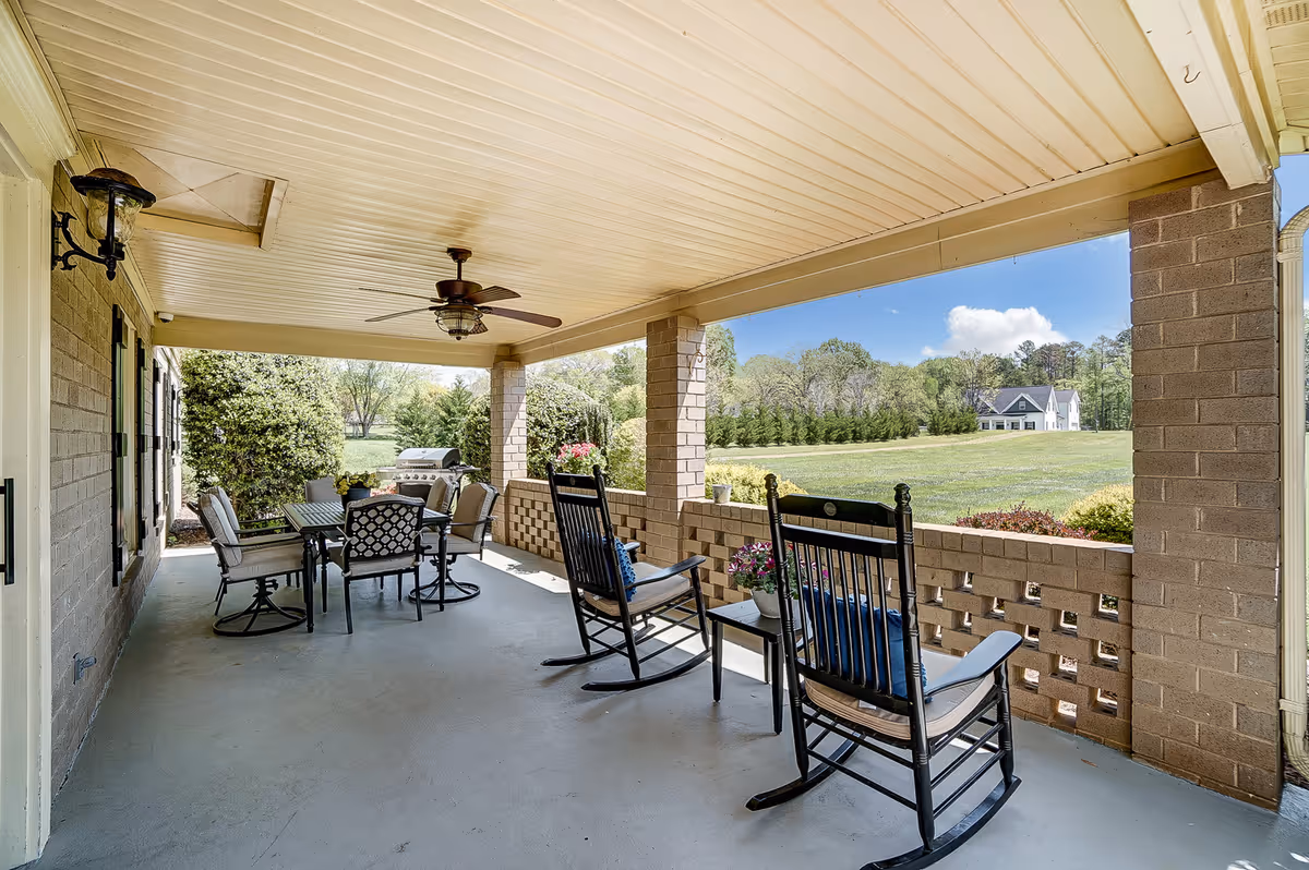 Covered outdoor patio with two black rocking chairs and a small table with potted flowers, a dining table with six cushioned chairs, a ceiling fan, and a grill. The patio overlooks a large grassy field with trees and a house in the distance under a blue sky with some clouds.