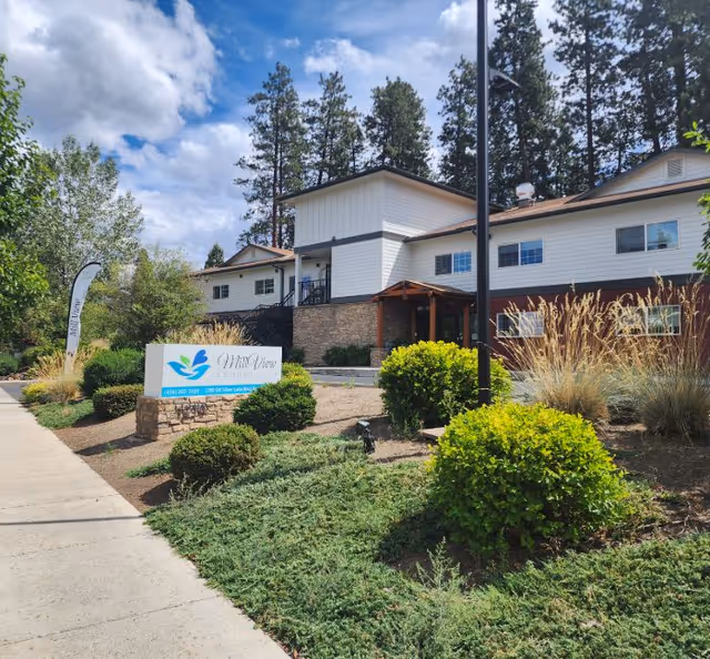 Exterior view of Mill View Memory Care facility showing a two-story building with white and brown siding, surrounded by landscaped bushes, trees, and a sidewalk under a partly cloudy sky.