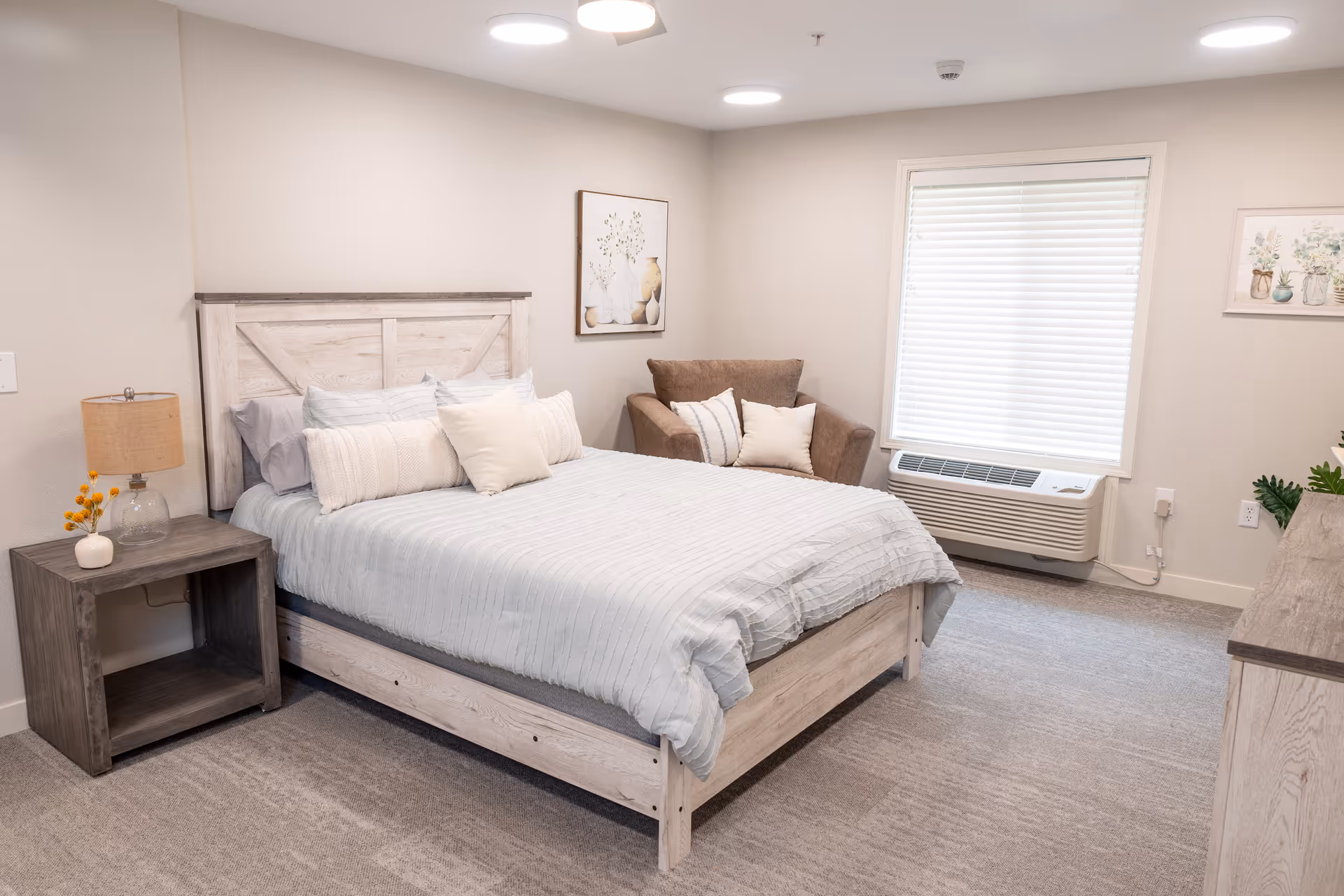A bright and cozy bedroom featuring a wooden bed with light gray bedding and multiple pillows. To the left of the bed is a wooden nightstand with a lamp and a small vase with yellow flowers. A brown armchair with cushions is placed near a window with white blinds. The room has light-colored walls, carpeted flooring, and framed artwork on the walls.