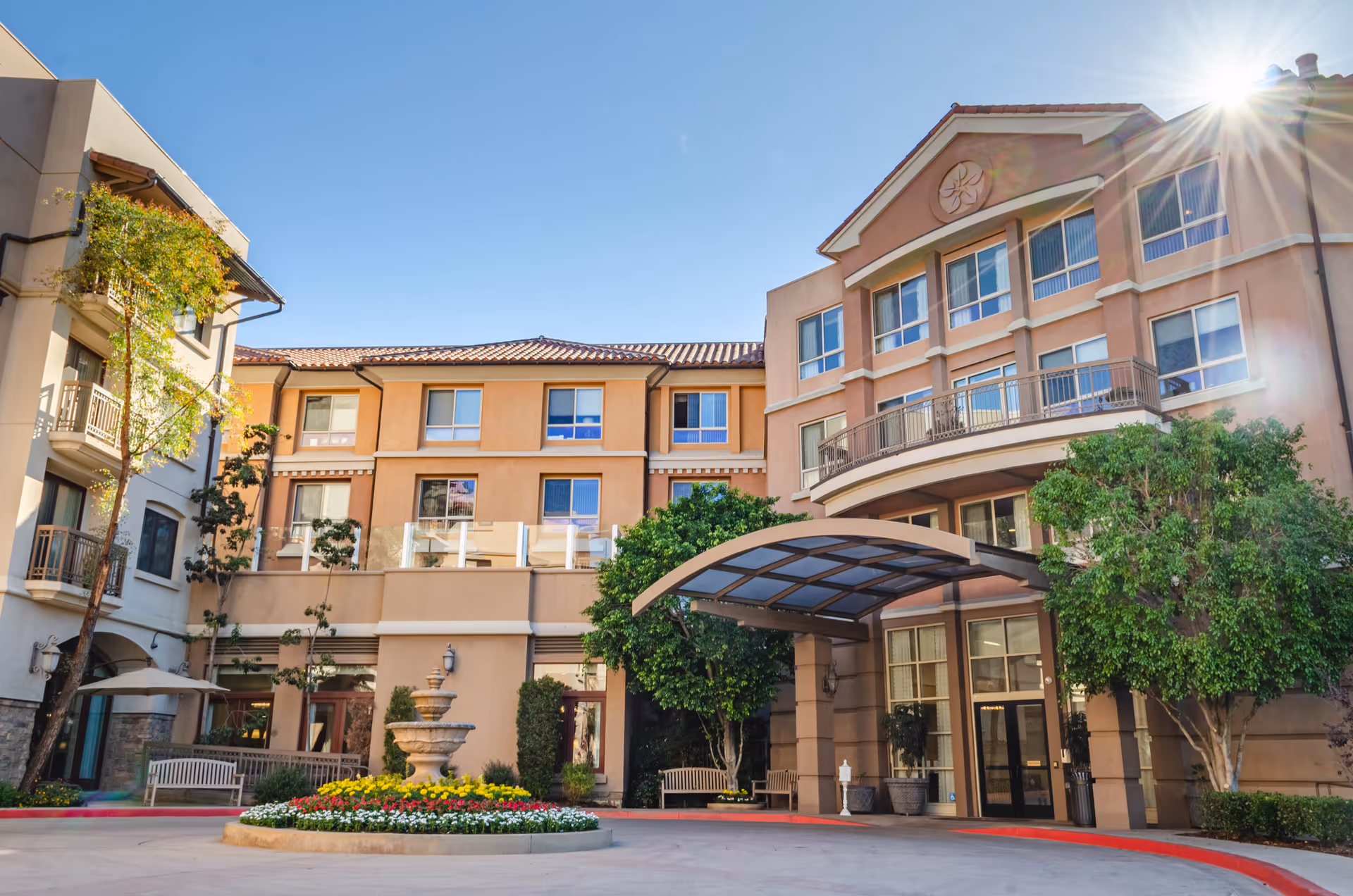 Sunny front entrance and courtyard of a multi-story senior living building with a covered porte-cochère, fountain, benches, and balconies.