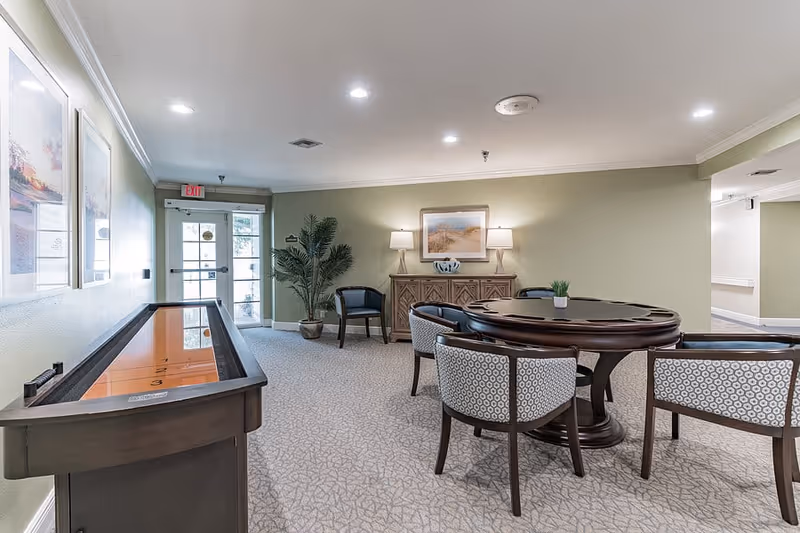 A well-lit common area in a senior living facility featuring a round wooden table with four patterned chairs around it, a shuffleboard table along the left wall, a wooden sideboard with two lamps and a framed picture above it, a potted plant, and a glass door exit in the background.