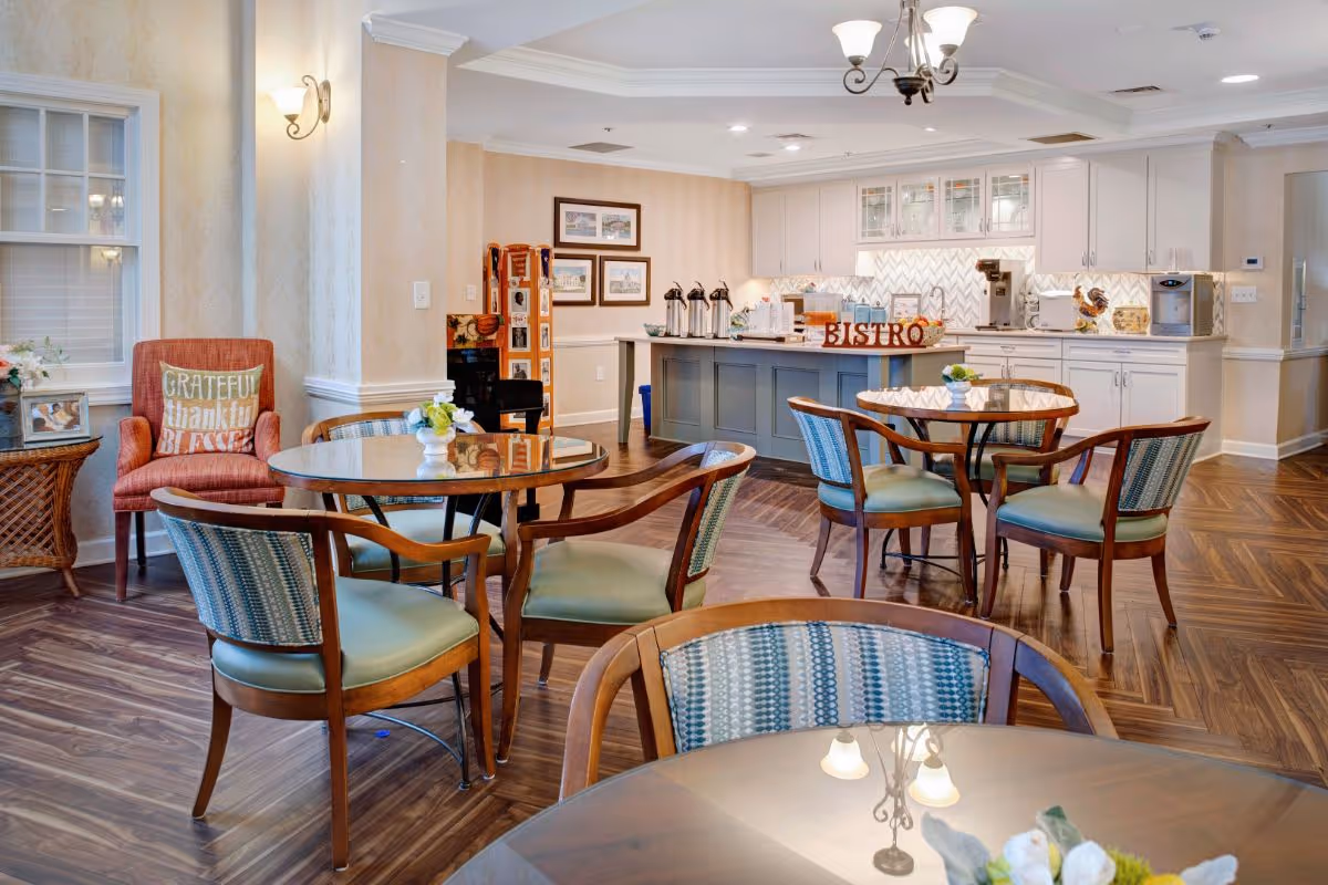 Bright communal bistro-style dining area with round tables and chairs in front of a serving counter and kitchenette.