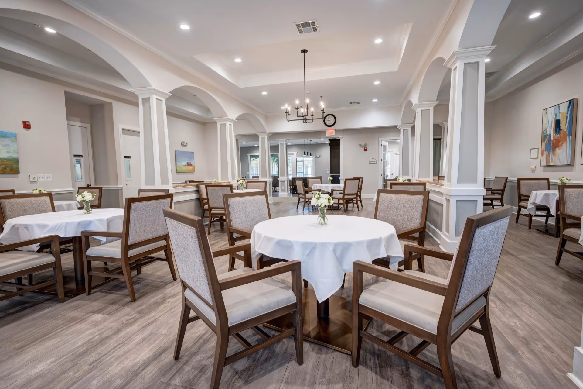 A spacious dining room in an assisted living facility with multiple round tables covered with white tablecloths, each surrounded by wooden chairs with cushioned seats and backs. The room features arched columns, light wood flooring, recessed lighting, and decorative wall art. Small flower arrangements are placed on each table.
