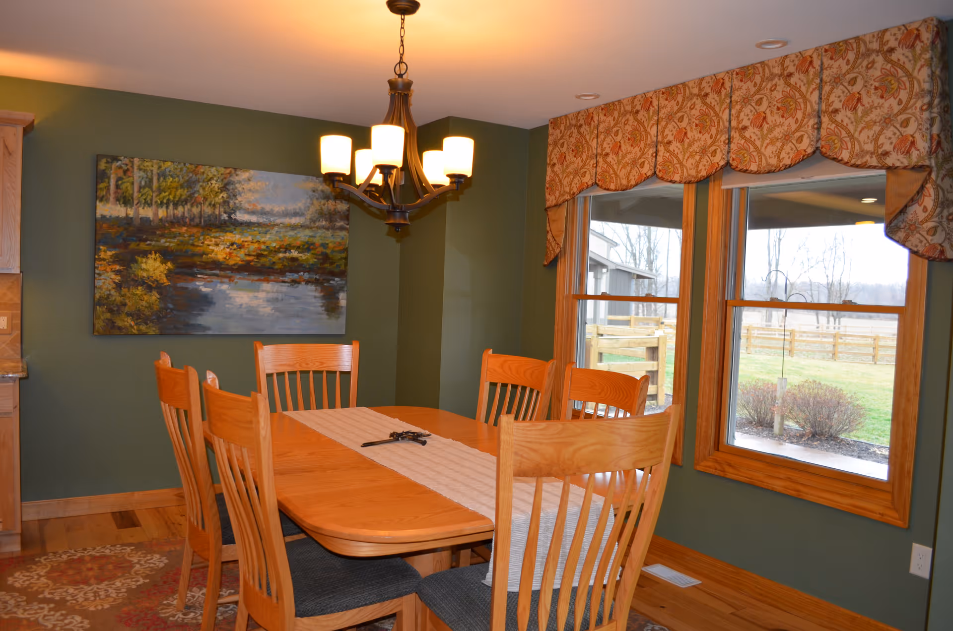 Dining room with a wooden table and chairs under a chandelier, green walls, a landscape painting, and windows overlooking a yard.