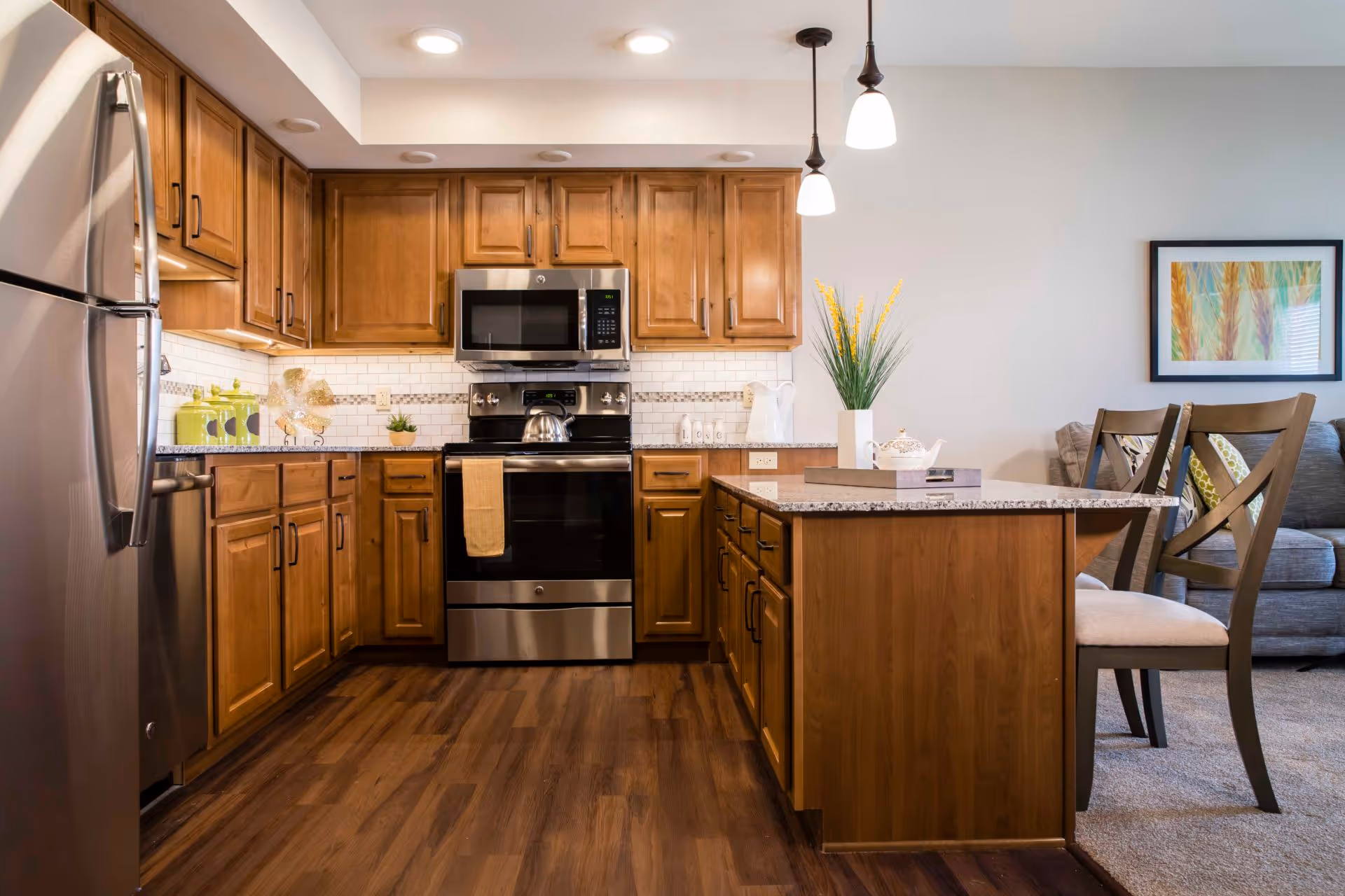 A modern kitchen with wooden cabinets, stainless steel refrigerator, oven, and microwave. The kitchen has a granite countertop island with two chairs, a vase with yellow flowers, and a teapot on a tray. The floor is wooden, and the kitchen opens into a living area with a gray sofa and a framed artwork on the wall.