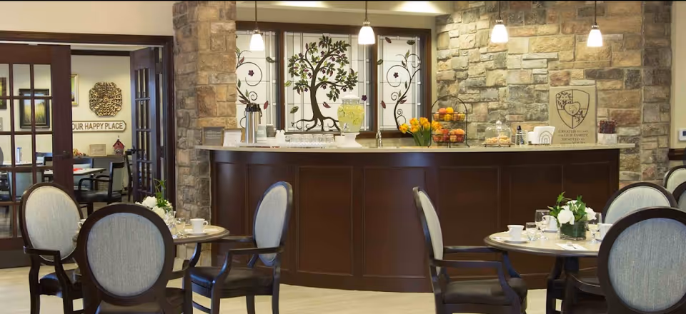 A cozy dining area with round tables set with cups, glasses, and small flower arrangements. The room features a stone wall with a counter displaying a beverage dispenser, fruit baskets, and pastries. Behind the counter are decorative stained glass windows with tree and floral designs. The space is softly lit with hanging pendant lights and has wooden framed chairs with cushioned seats.
