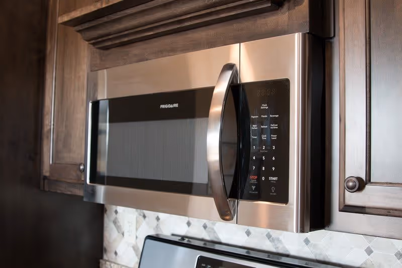 Close-up view of a stainless steel Frigidaire microwave oven mounted above a stove, surrounded by dark wooden kitchen cabinets and a tiled backsplash.