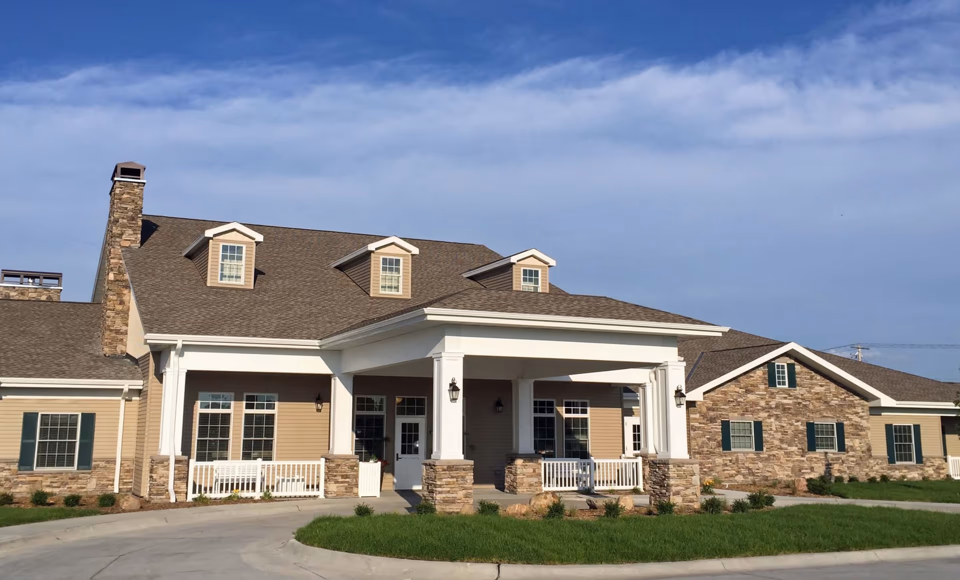 Front view of a residence building with a covered entrance portico, stone accents, dormer windows, and a manicured lawn under a blue sky.