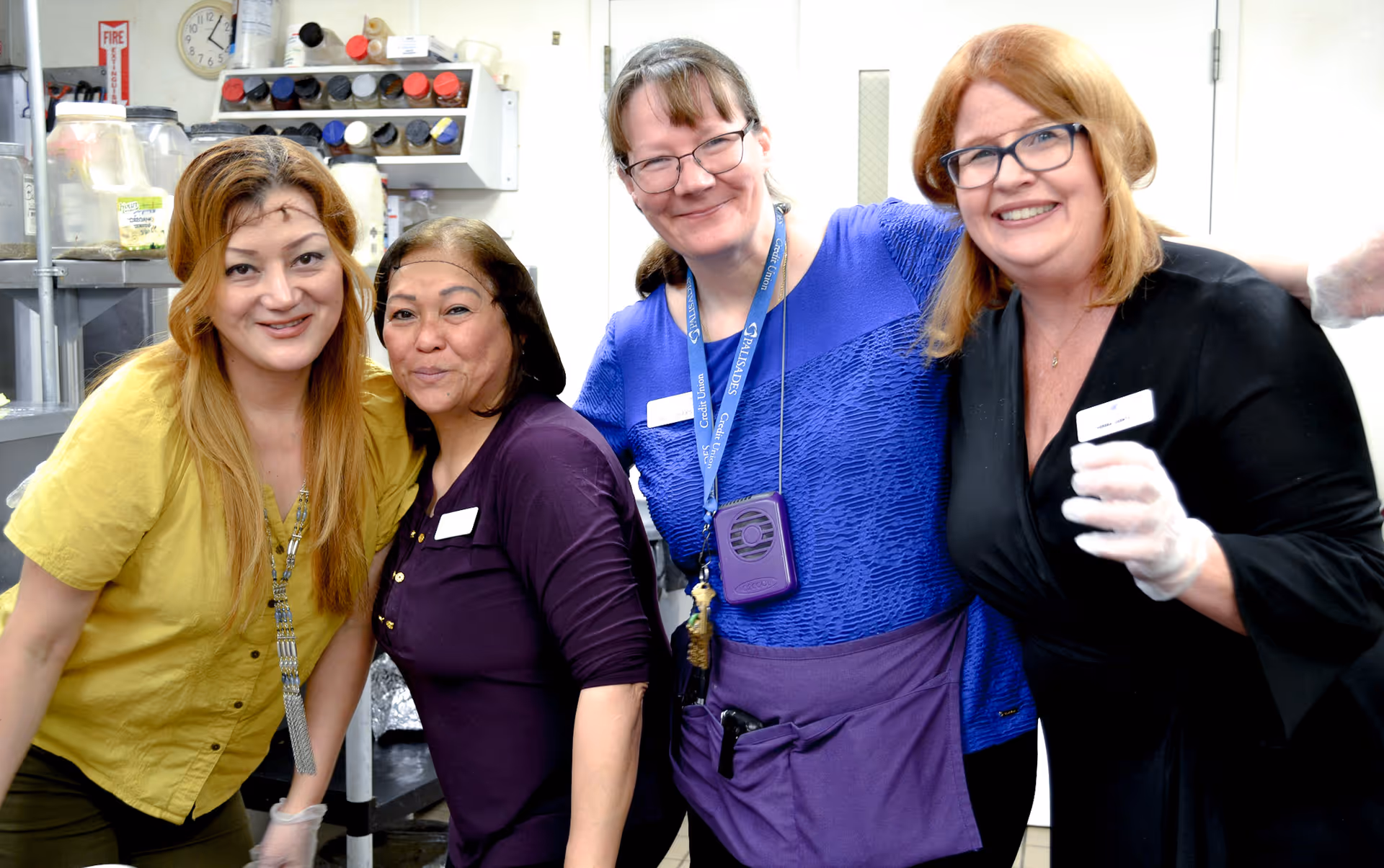 Four women smiling and posing together in a kitchen setting. They are wearing name tags and some have gloves on, standing close to each other with kitchen equipment and shelves with containers in the background.