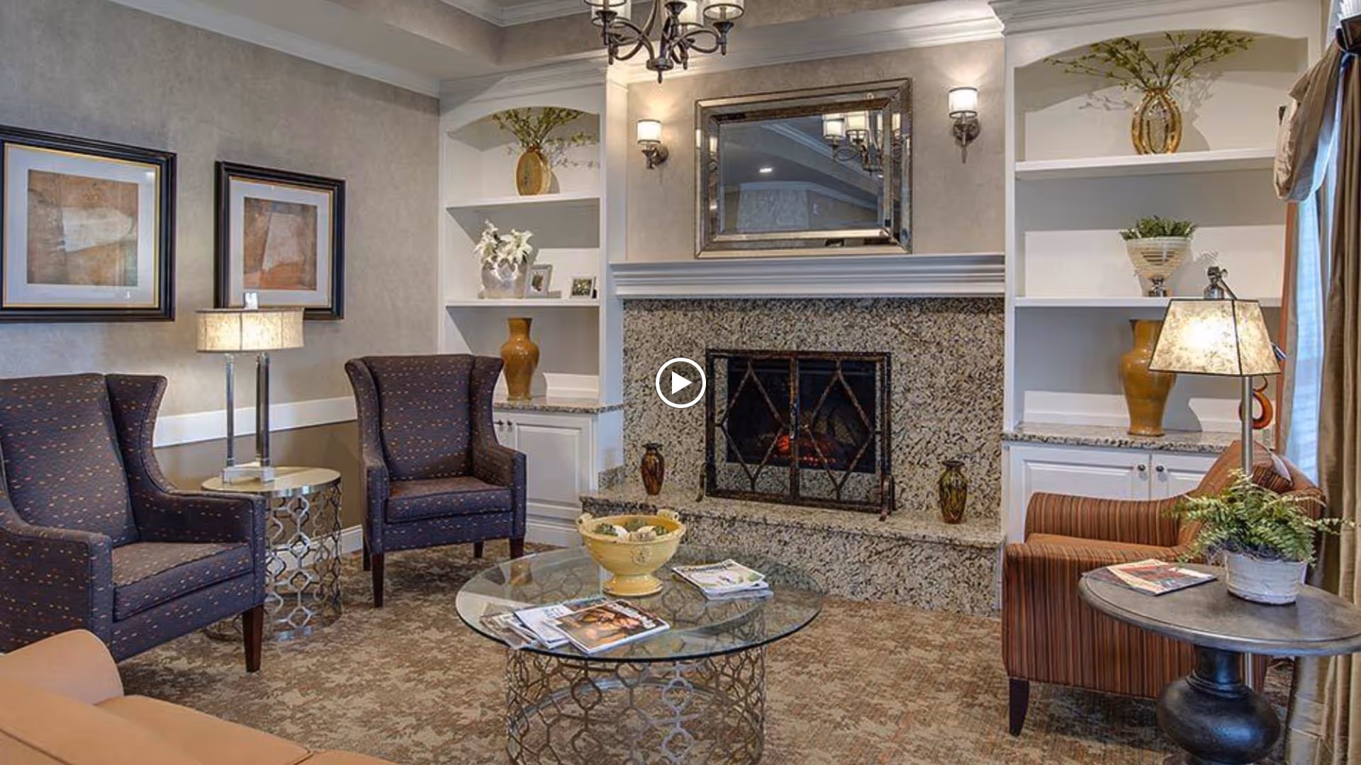 A cozy senior living room with a granite fireplace in the center, flanked by built-in white shelves decorated with vases and plants. There are two dark upholstered armchairs with a small round side table and lamp between them on the left, and a striped armchair with a round side table and lamp on the right. A glass coffee table with magazines and a decorative bowl sits in the middle on a patterned carpet. Two framed abstract artworks hang on the left wall.