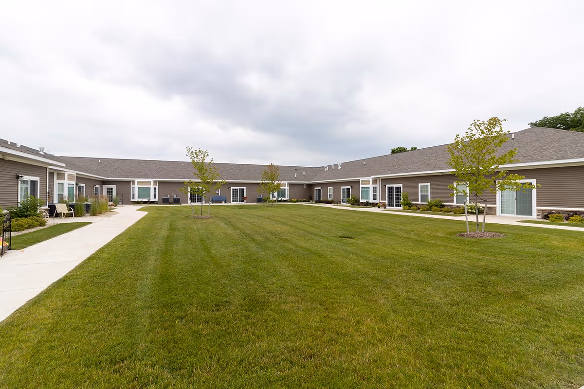 A large, well-maintained grassy courtyard surrounded by a single-story building with multiple windows and sliding glass doors. There are small trees planted in the grass and a concrete walkway along the building's perimeter. The sky is overcast.