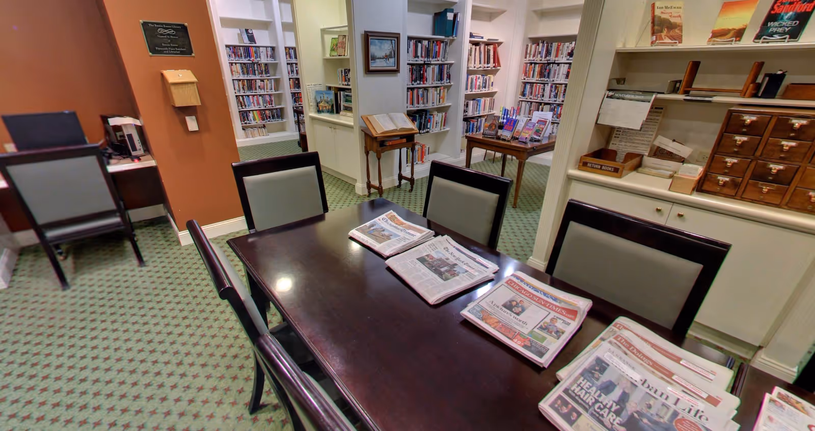 Interior view of a library room at Plymouth Place featuring a dark wooden table with newspapers spread out on top, surrounded by chairs with light-colored cushions. Bookshelves filled with books line the walls, and there is a small wooden stand with an open book. The carpet has a green pattern, and there is a plaque on the wall along with a small desk and chair in the corner.