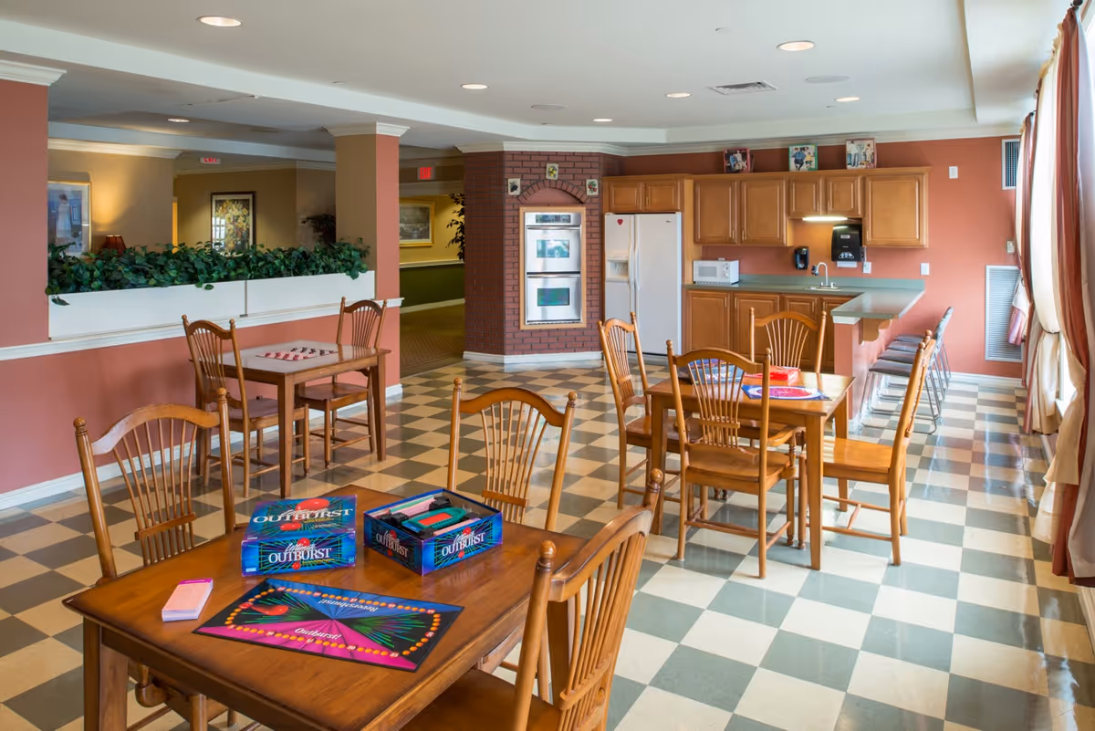 A bright and spacious common area in an assisted living facility featuring a checkered floor, wooden tables and chairs, a kitchen area with wooden cabinets, a refrigerator, microwave, and built-in ovens. Board games are set up on one of the tables, and large windows with curtains allow natural light to fill the room.