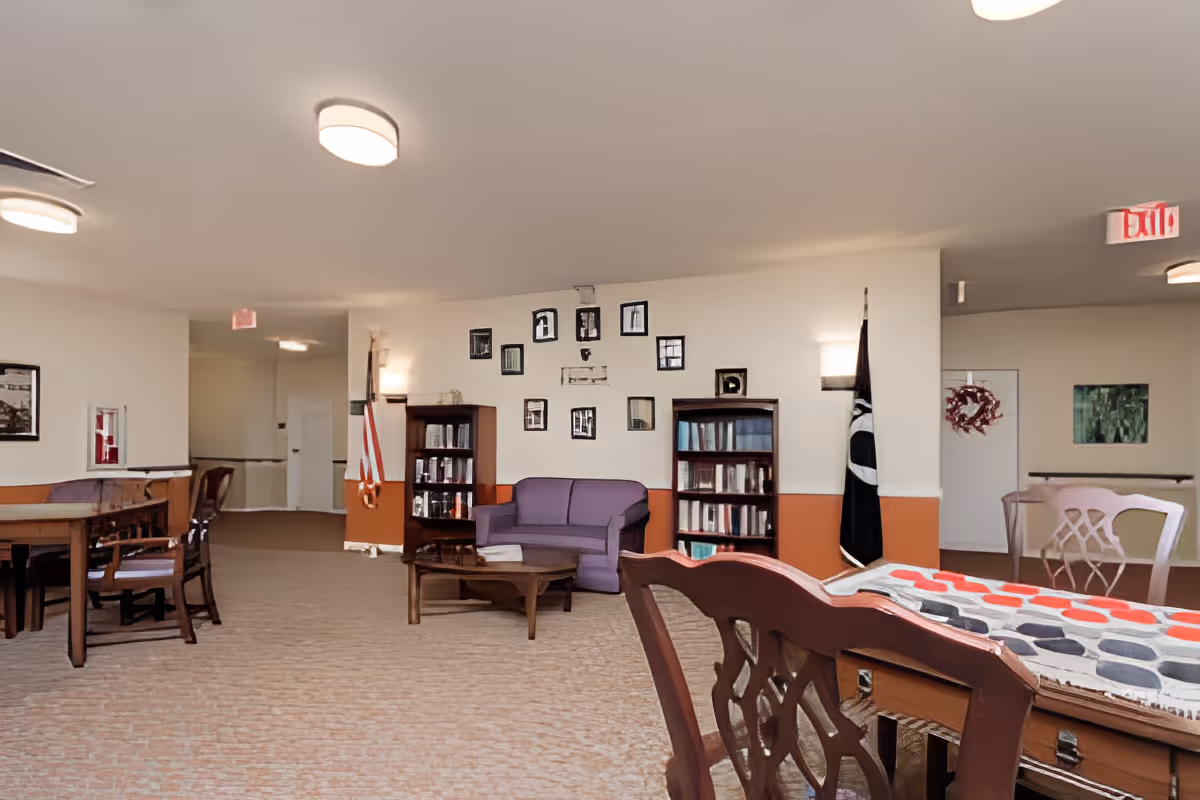A spacious common area in a senior living facility with carpeted floors, several wooden tables and chairs, a purple loveseat, two bookshelves filled with books, framed pictures on the wall, and flags displayed near the walls. The room is well-lit with ceiling lights and wall sconces, and there is an exit sign visible in the background.
