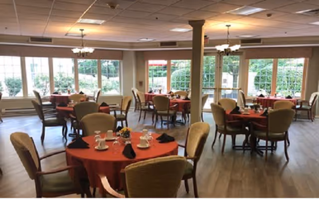 Spacious dining room with round tables covered in red tablecloths, place settings and upholstered chairs beneath chandeliers by large windows.