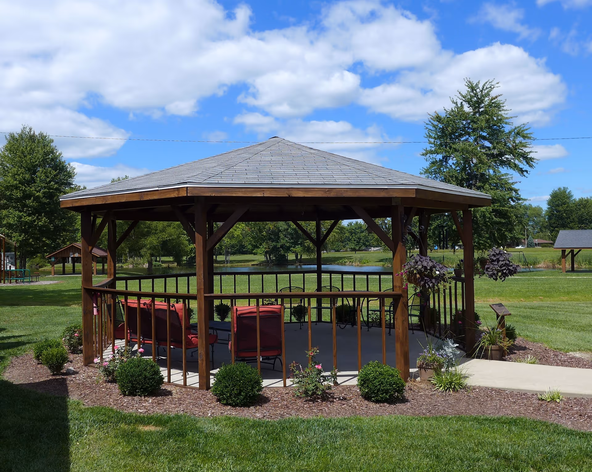 Wooden gazebo with red cushioned chairs on a concrete pad in a landscaped grassy area overlooking a pond under a blue sky.