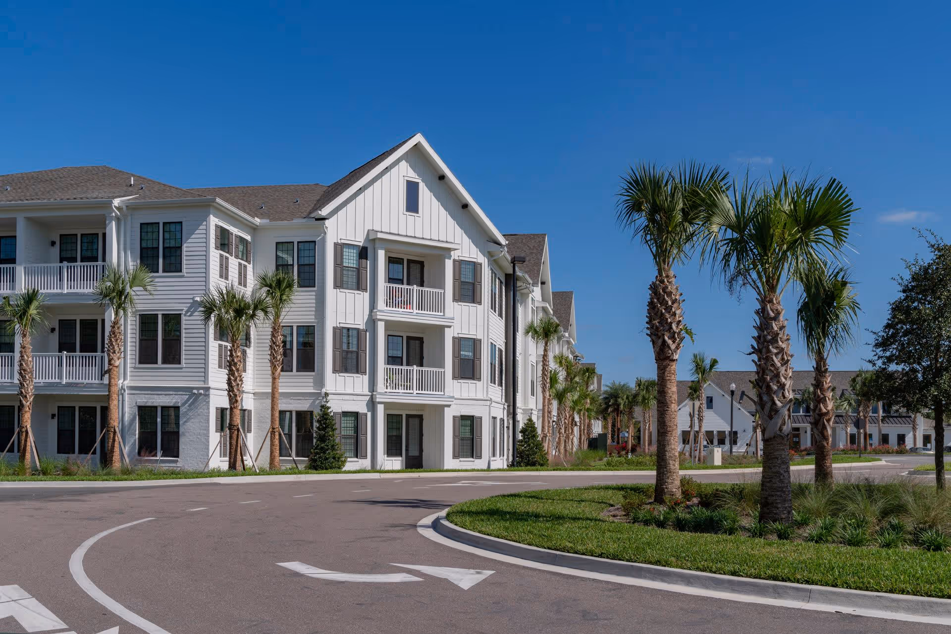 Exterior view of a modern senior living facility with white siding and multiple balconies, surrounded by palm trees and a curved driveway under a clear blue sky.