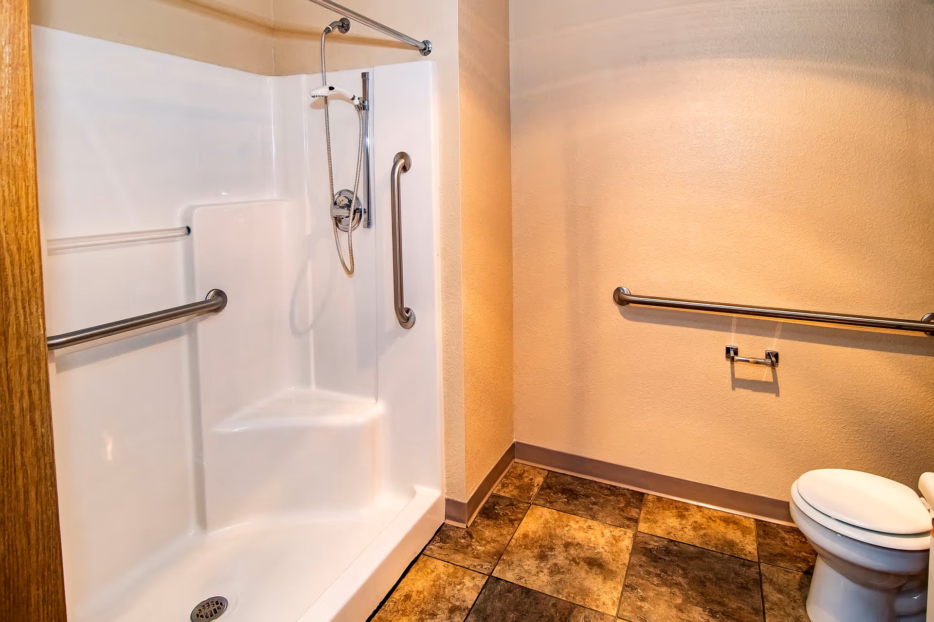 A bathroom with a white walk-in shower featuring a built-in seat and grab bars, a toilet with a grab bar beside it, and tiled flooring in shades of brown and beige.