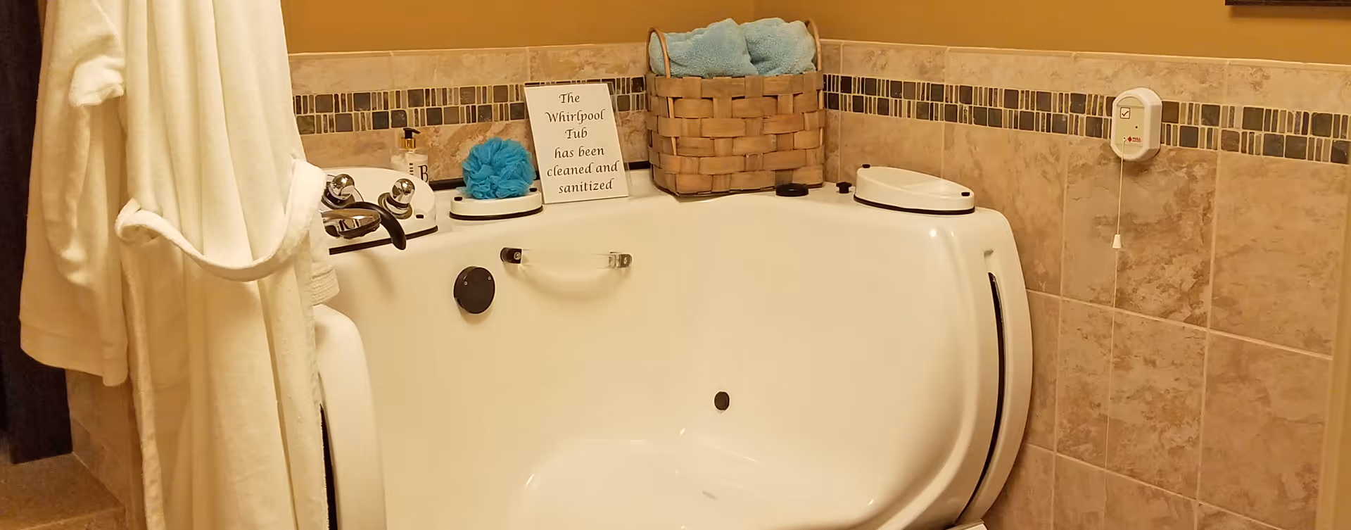 A clean white whirlpool bathtub in a bathroom with beige tiled walls and a decorative tile border. A white bathrobe hangs on the left side. On the edge of the tub, there is a basket with blue towels, a blue bath sponge, a soap dispenser, and a sign that reads 'The Whirlpool Tub has been cleaned and sanitized.' A pull cord emergency alert device is mounted on the wall to the right.