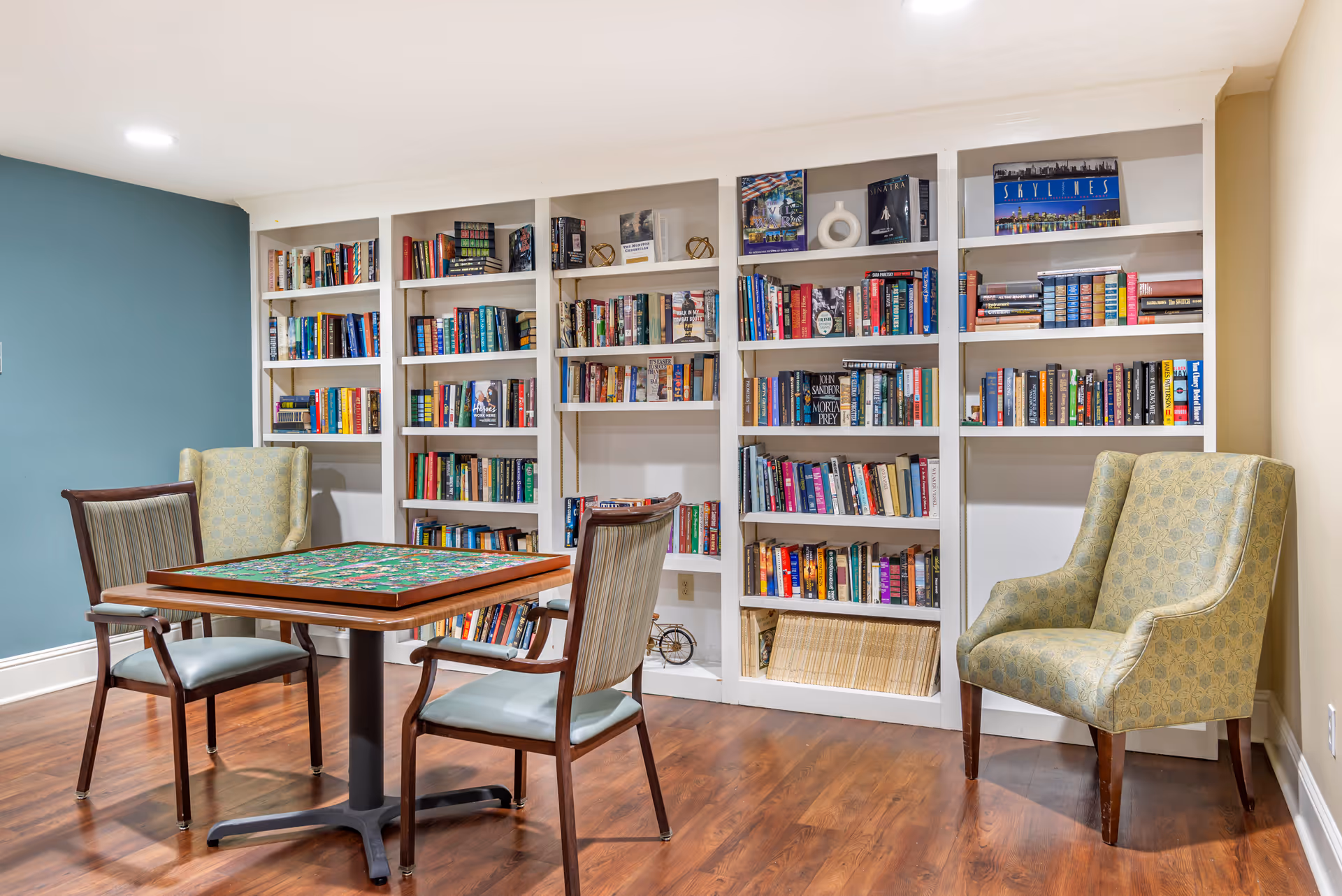 A cozy reading and game area in a senior living facility featuring a wooden table with a puzzle on it, surrounded by four chairs. Behind the table is a large built-in bookshelf filled with various books and decorative items. The room has wooden flooring and walls painted in light beige and teal colors.