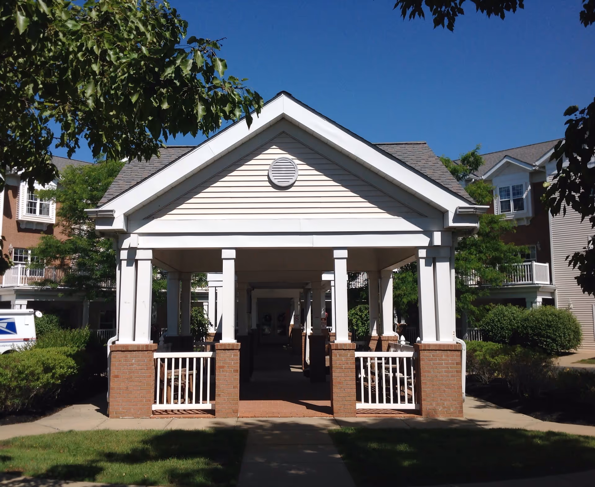 Covered walkway with white columns and brick bases leading through a garden area with green bushes and trees, under a clear blue sky. Residential-style buildings are visible in the background.