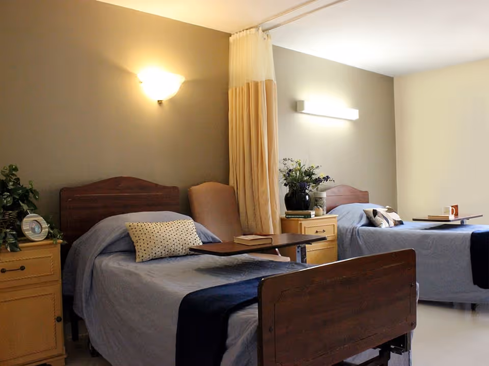 A room with two single beds each with blue bedding and a pillow. Between the beds is a beige curtain divider, a beige armchair, and a nightstand with a vase of flowers and books. Each bed has an overbed table, one with a book and the other with a cup. The walls are painted in neutral tones with wall-mounted lights above each bed.