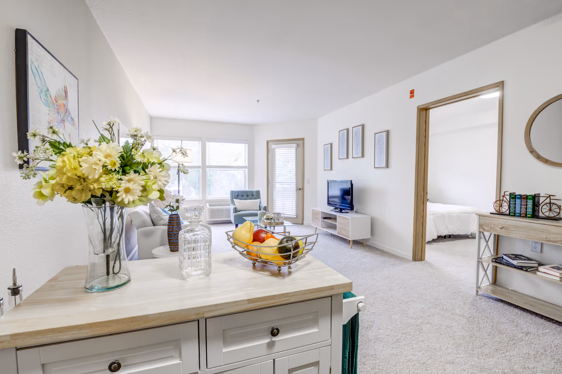 Bright and airy living room area with a light-colored sofa, a blue armchair, a TV on a white stand, and a glass coffee table. A wooden cabinet with a vase of flowers and a glass decanter is in the foreground. The room has large windows letting in natural light and an open doorway leading to a bedroom with a white bedspread. The walls are decorated with framed artwork and a round mirror above a console table with books and decorative items.