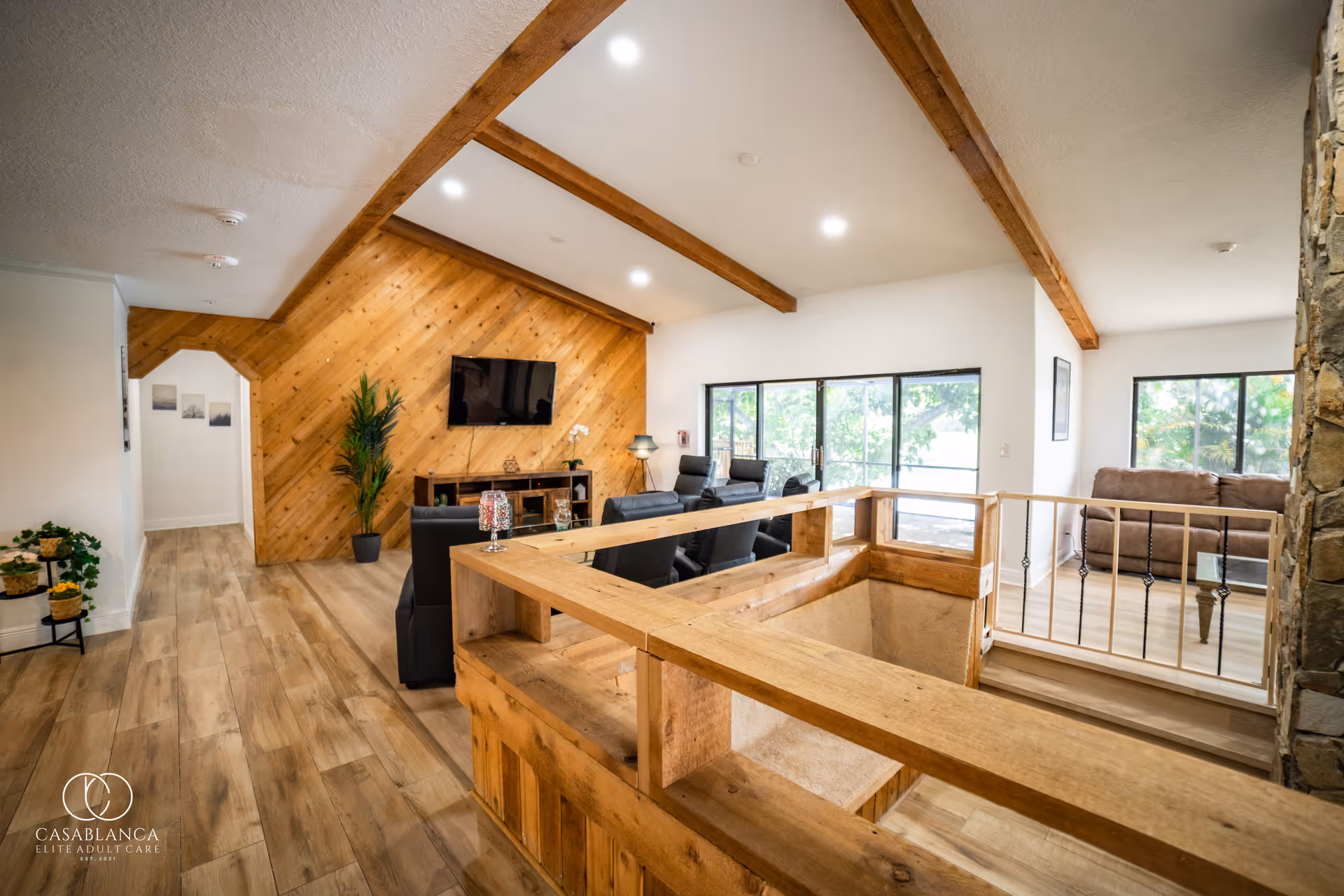 Spacious living room area with wooden beams on the ceiling and wood-paneled accent wall with a mounted flat-screen TV. The room features black leather recliners, large windows letting in natural light, and a wooden railing around a stairwell. There is a stone fireplace on the right and a brown sofa near the windows.