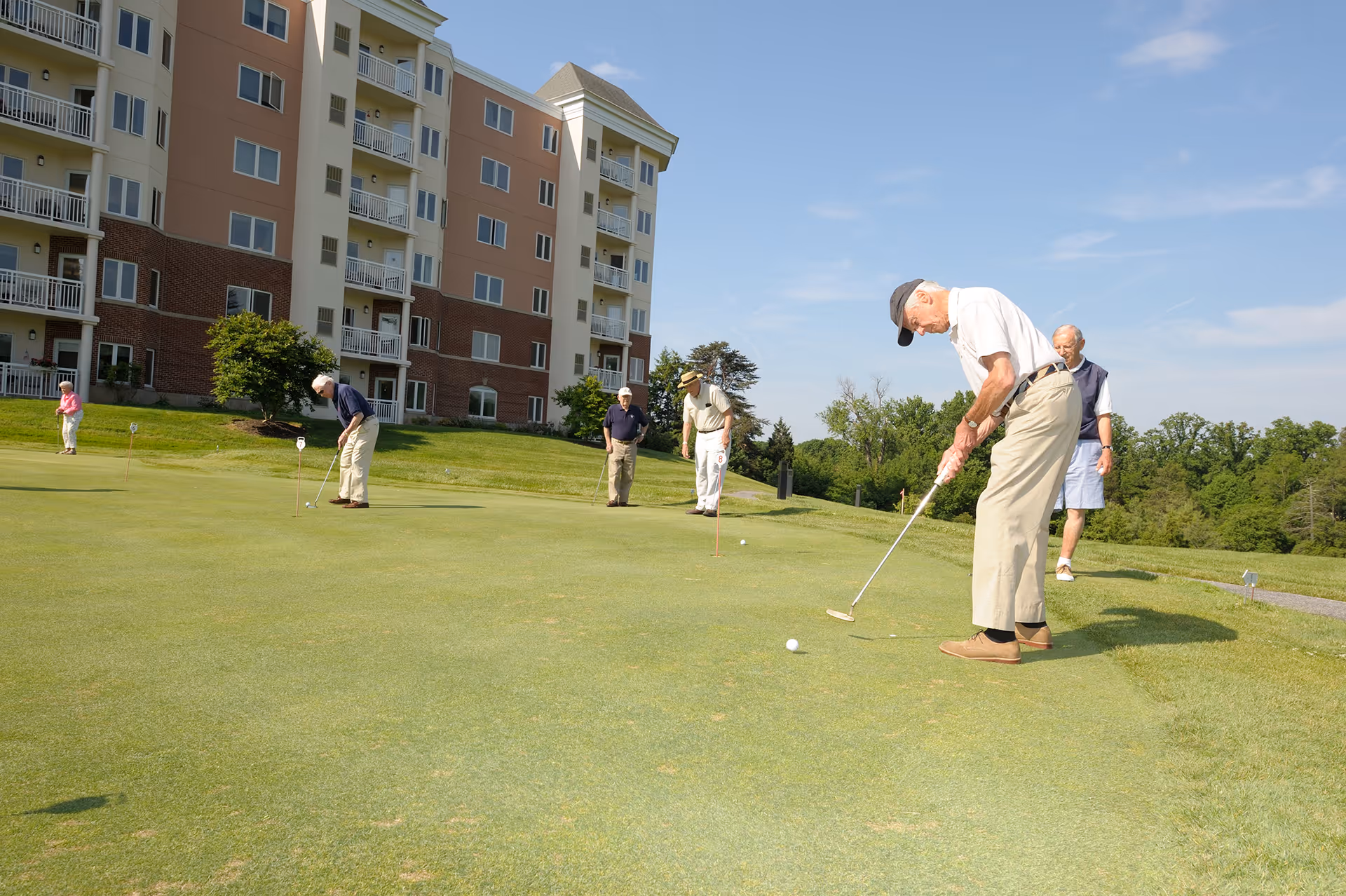 Several elderly people playing golf on a putting green outside a multi-story residential building under a clear blue sky.