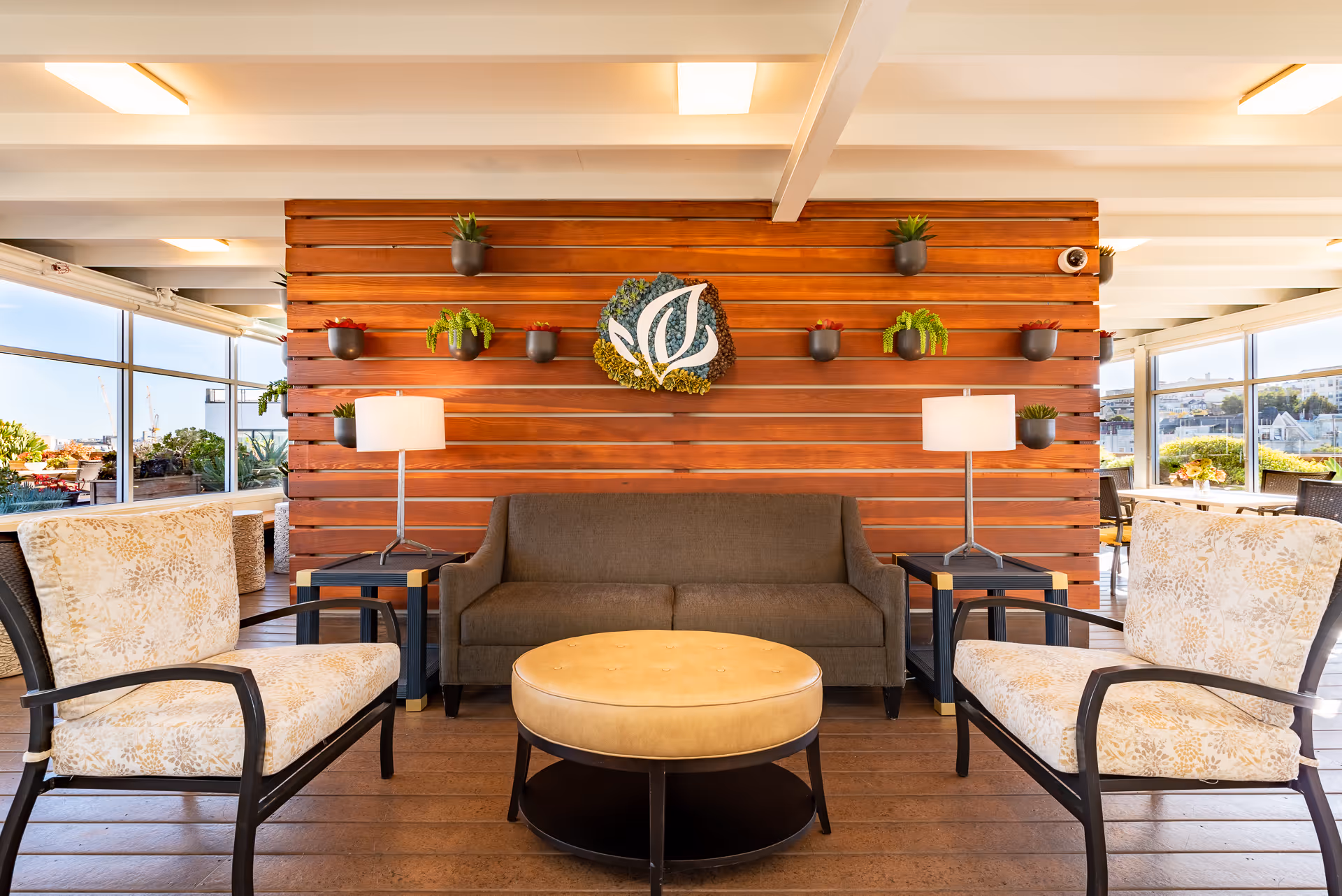 A cozy indoor seating area with a brown sofa, two floral-patterned armchairs, and a round beige ottoman coffee table. The background features a wooden slat wall decorated with small potted plants and a leafy logo in the center. Two side tables with lamps flank the sofa. Large windows on either side provide a view of an outdoor patio with additional seating and greenery.