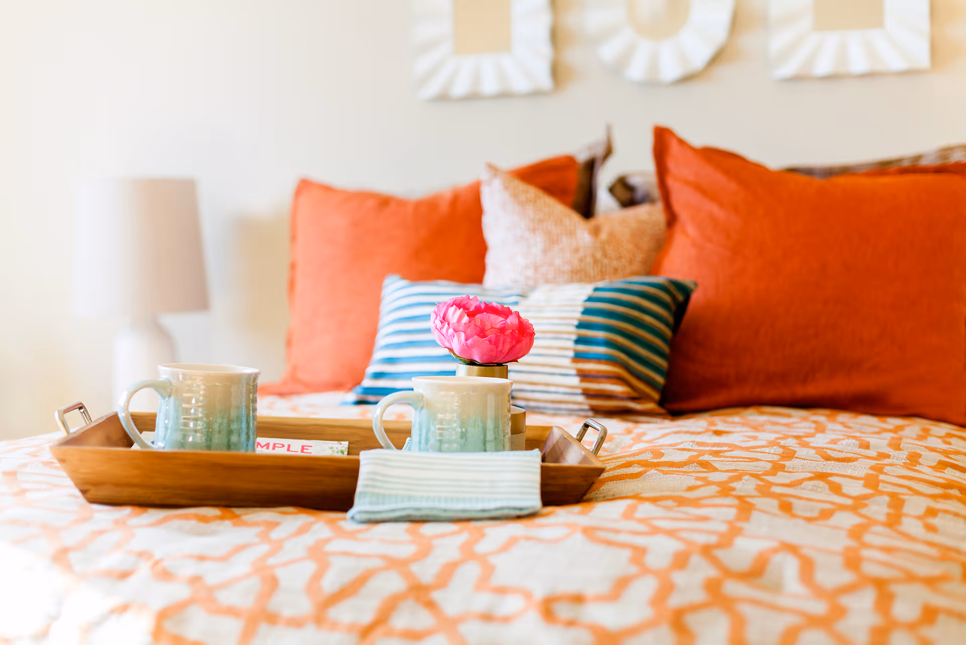A cozy bed with an orange and white patterned bedspread, decorated with multiple pillows in shades of orange, beige, and blue stripes. On the bed is a wooden tray holding two ceramic mugs, a small pink flower in a vase, and a folded cloth napkin. A white lamp is visible in the background.