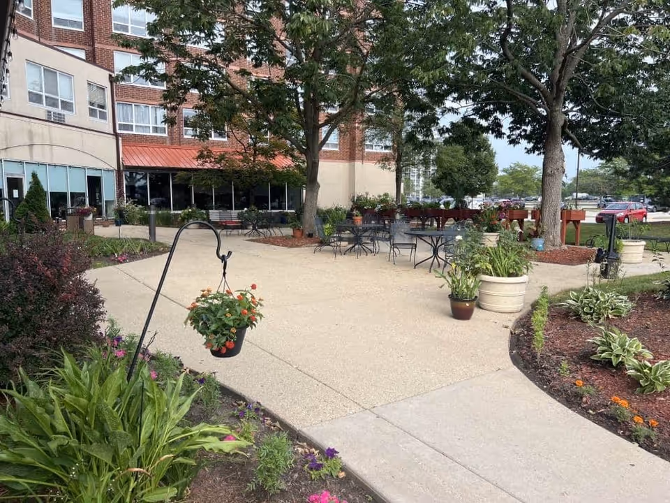 Outdoor patio area at Elison Independent Living of Niles with metal tables and chairs, surrounded by trees, potted plants, and flower beds. The building's brick exterior is visible in the background along with a parking lot and cars.