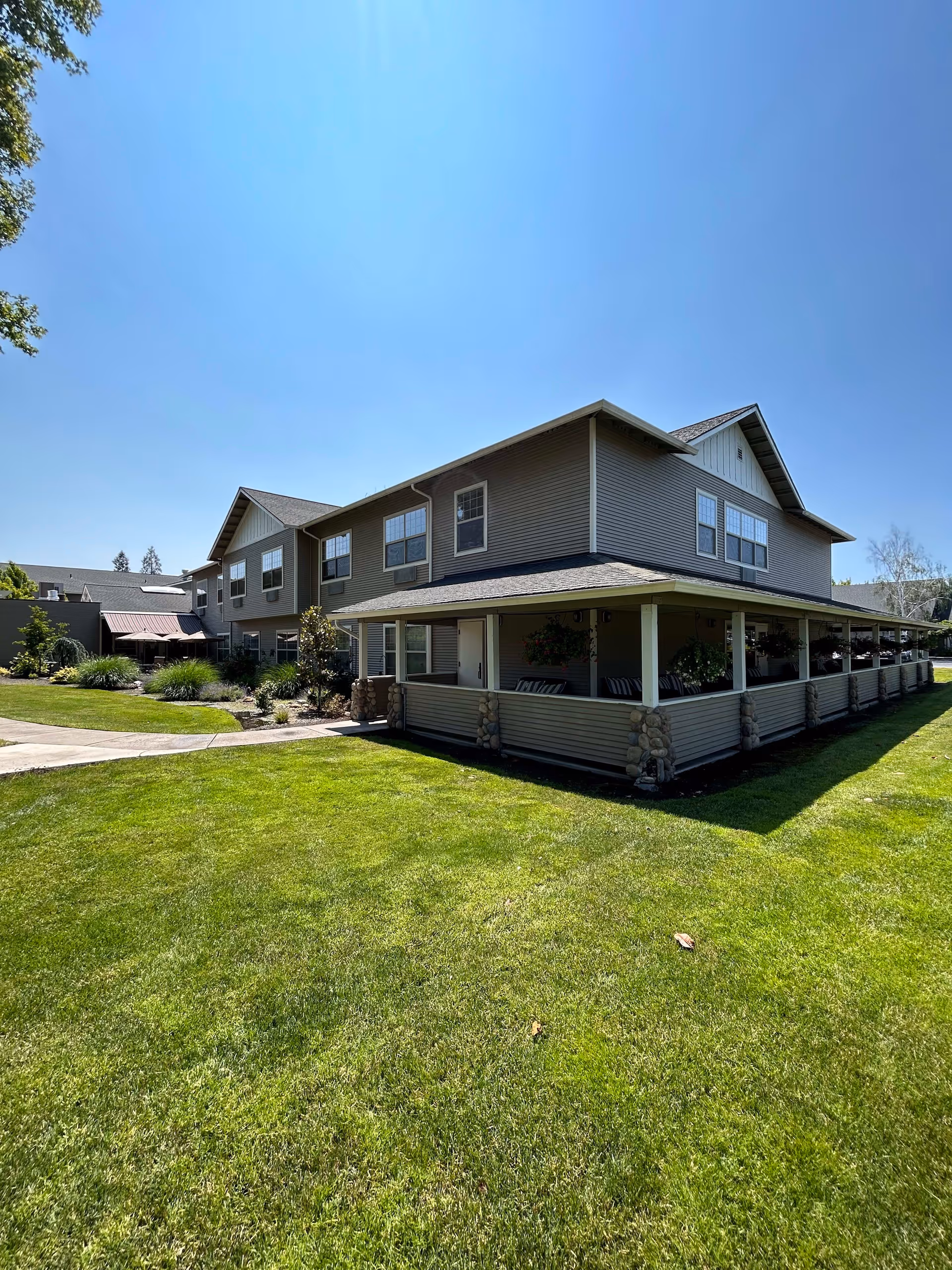 Exterior view of a two-story senior living facility building with beige siding and multiple windows. The building has a covered porch with stone pillars and hanging plants. The foreground features a well-maintained green lawn under a clear blue sky.