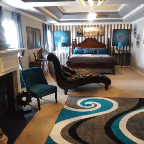 A cozy bedroom with a large wooden bed adorned with blue and brown pillows, including one with the word 'LOVE'. The room features a dark brown chaise lounge, a teal upholstered chair, a fireplace on the left, and a colorful area rug with swirling patterns in teal, black, white, and gray. The walls are decorated with framed artwork and two matching blue lamps on either side of the bed. The ceiling has a tray design with recessed lighting.